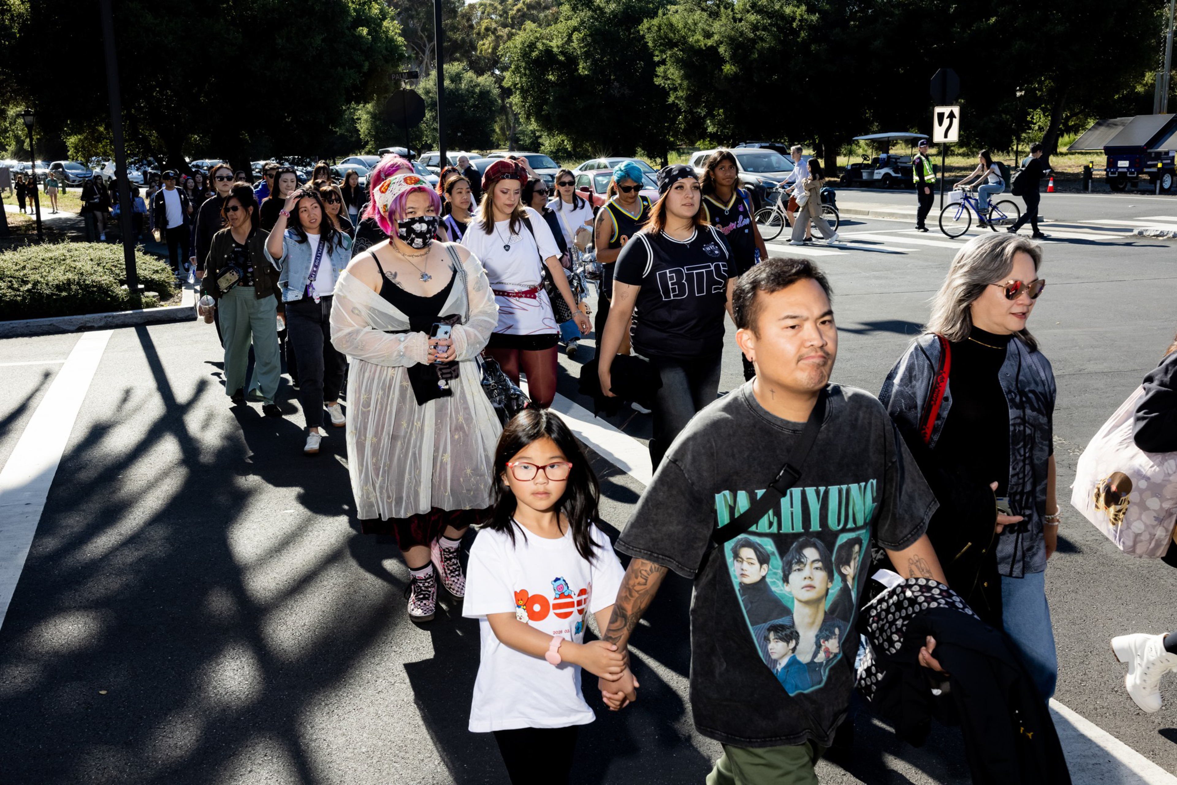 A diverse group of people, including adults and children, walk together across a street on a sunny day, some wearing graphic shirts and casual clothing.
