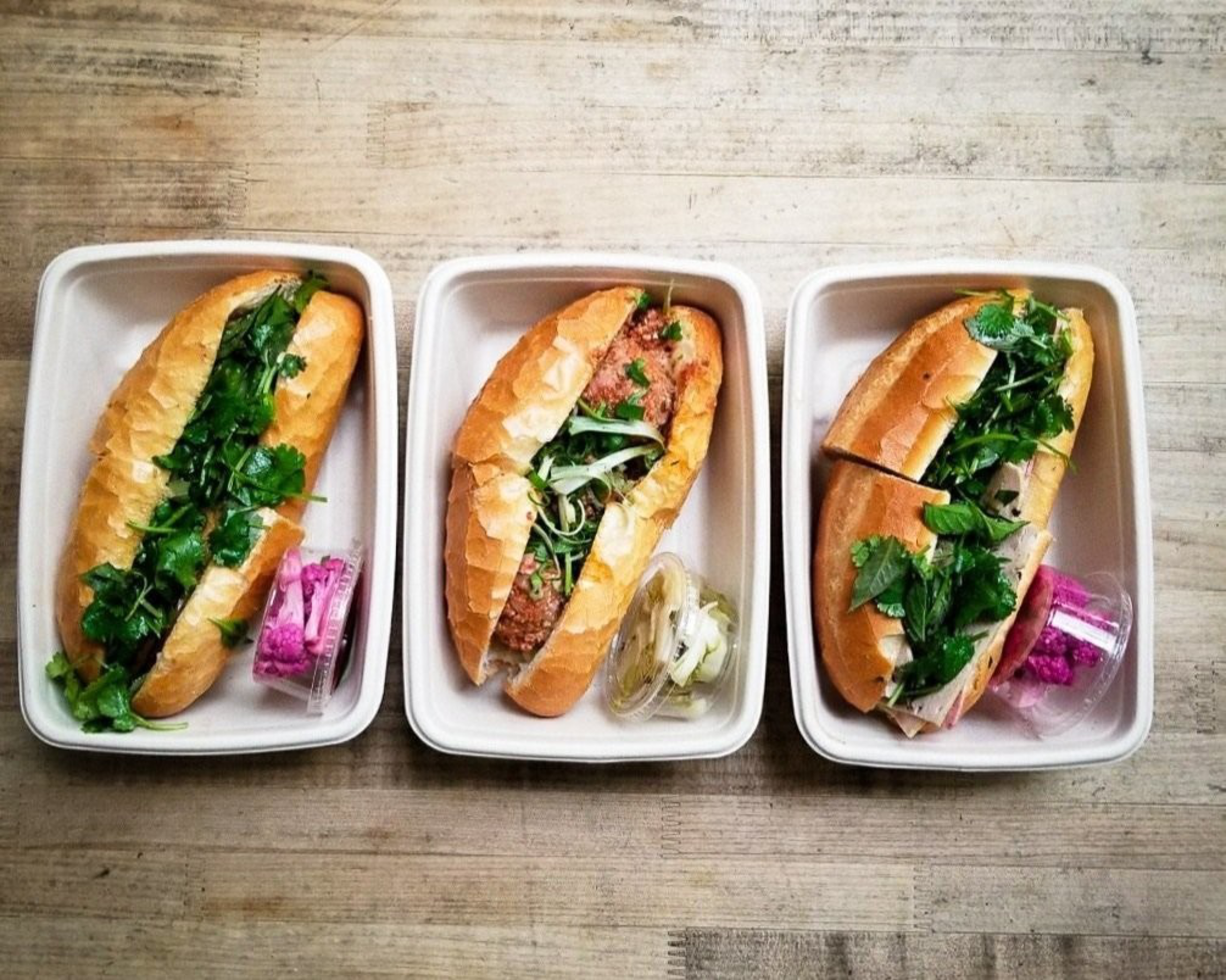 Three sandwiches filled with greens are placed in separate white containers, each accompanied by a small container of pickled vegetables.