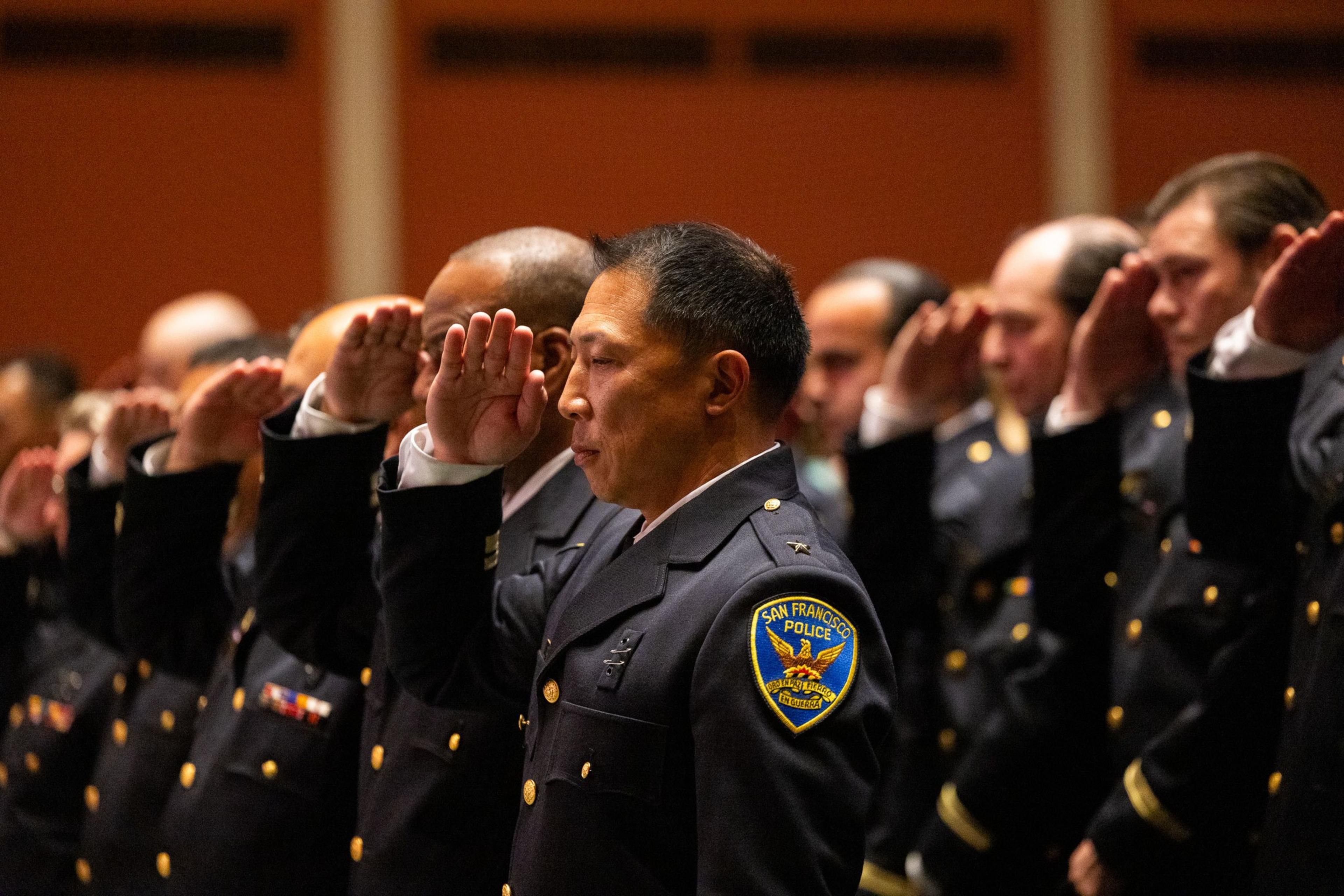 Uniformed San Francisco police officers stand in a row, saluting with their right hands raised to their foreheads during a formal ceremony.