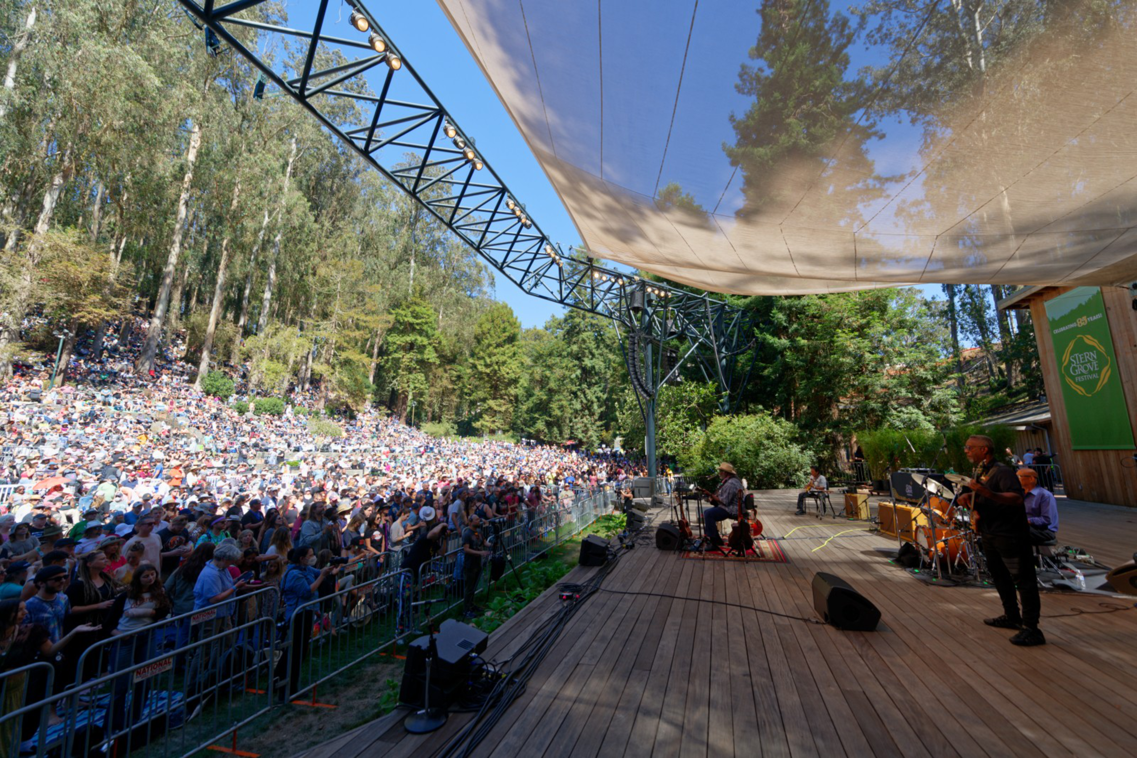 A large crowd watches a band performing on an outdoor wooded stage under a canopy on a sunny day.