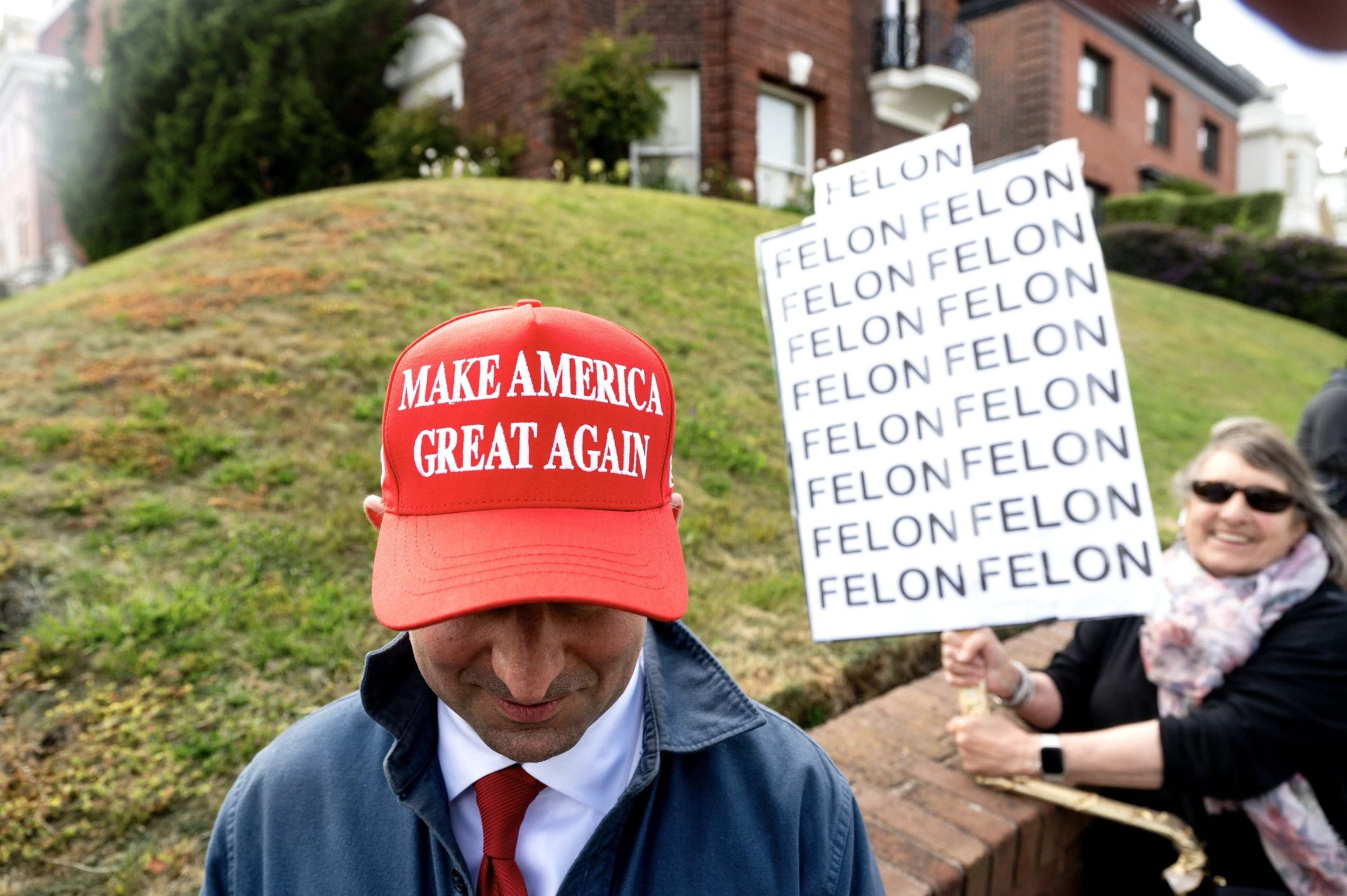 A person wearing a red &quot;Make America Great Again&quot; hat is looking down, while behind them, another person holds a sign repeatedly saying &quot;FELON.&quot;