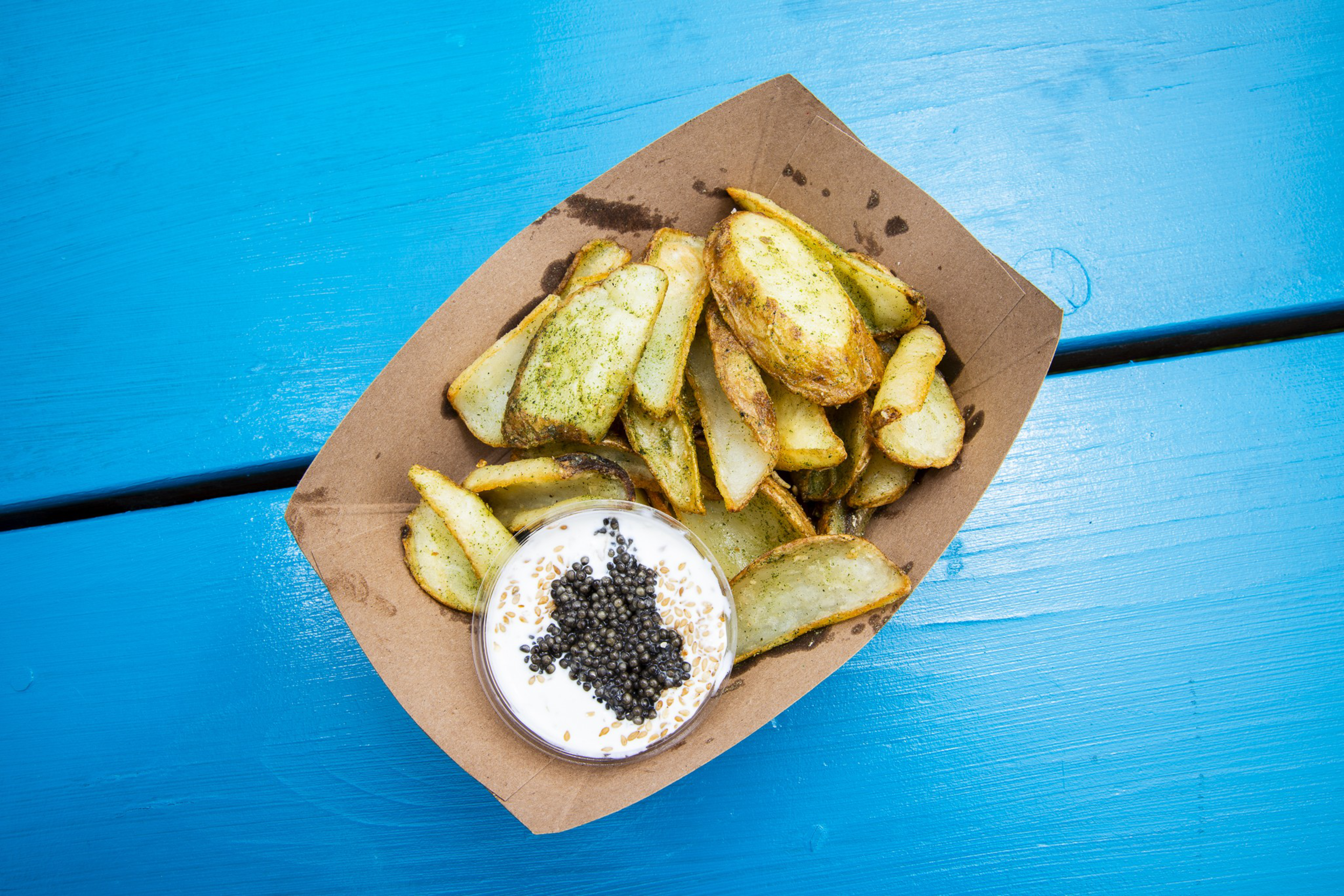 Golden potato wedges seasoned with herbs are served in a brown paper tray alongside a creamy dip topped with black caviar, all on a bright blue wooden surface.