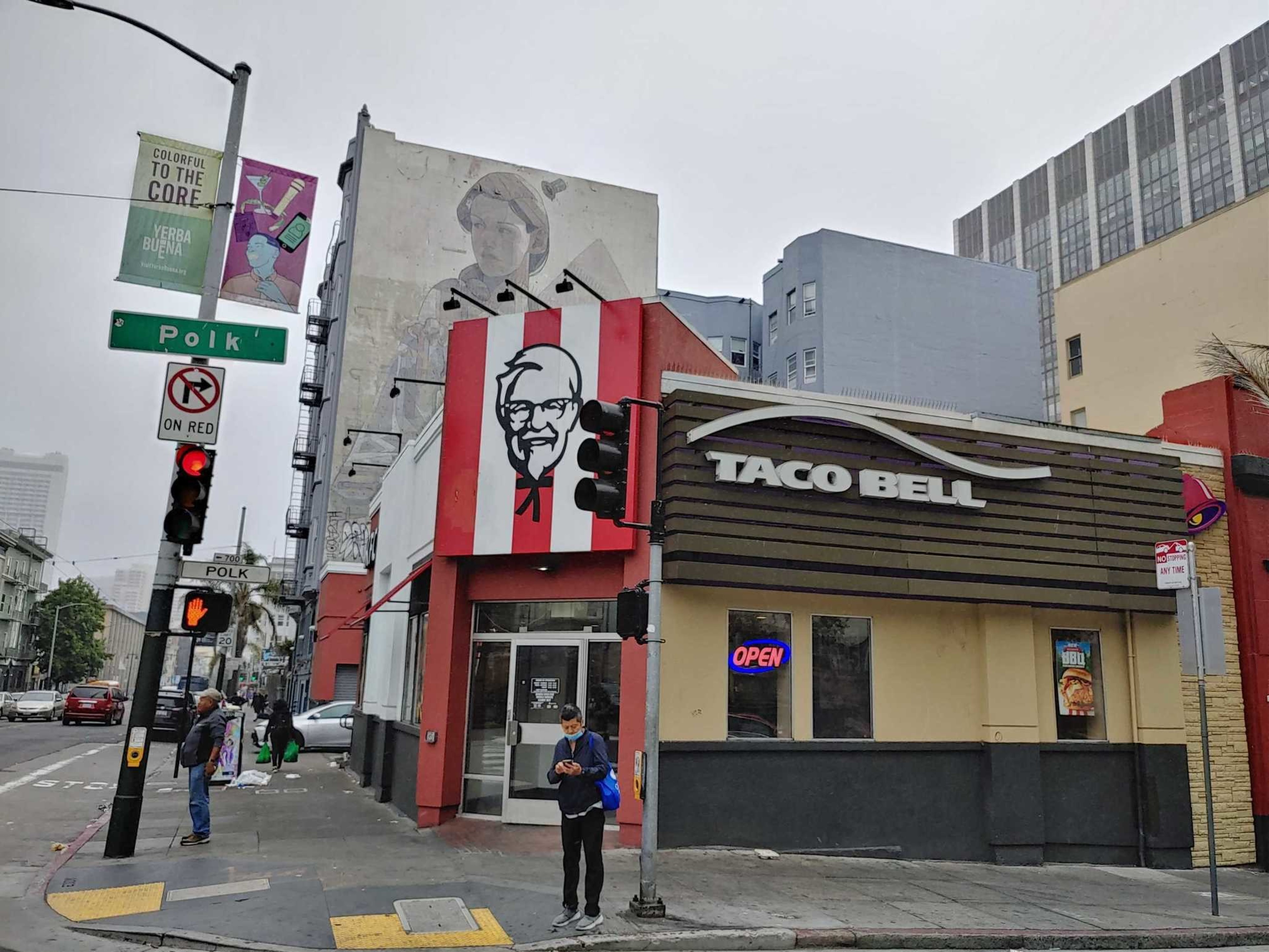 A KFC and Taco Bell share a corner building at the intersection of Polk Street, with people waiting at a crosswalk and a mural on a nearby wall.