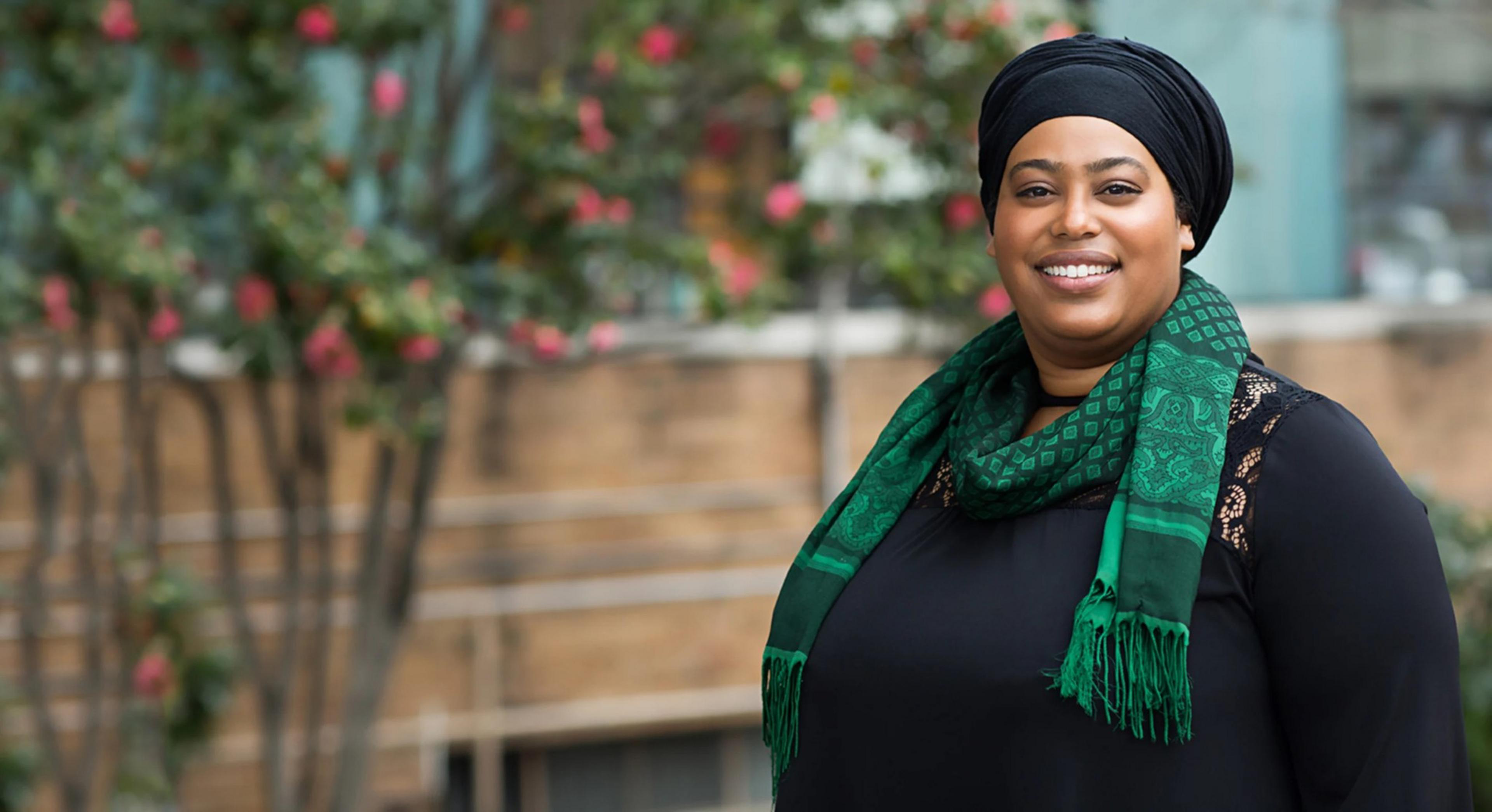 A smiling individual wearing a black top, black headwrap, and a green patterned scarf poses in front of a blurred background with trees and flowers.