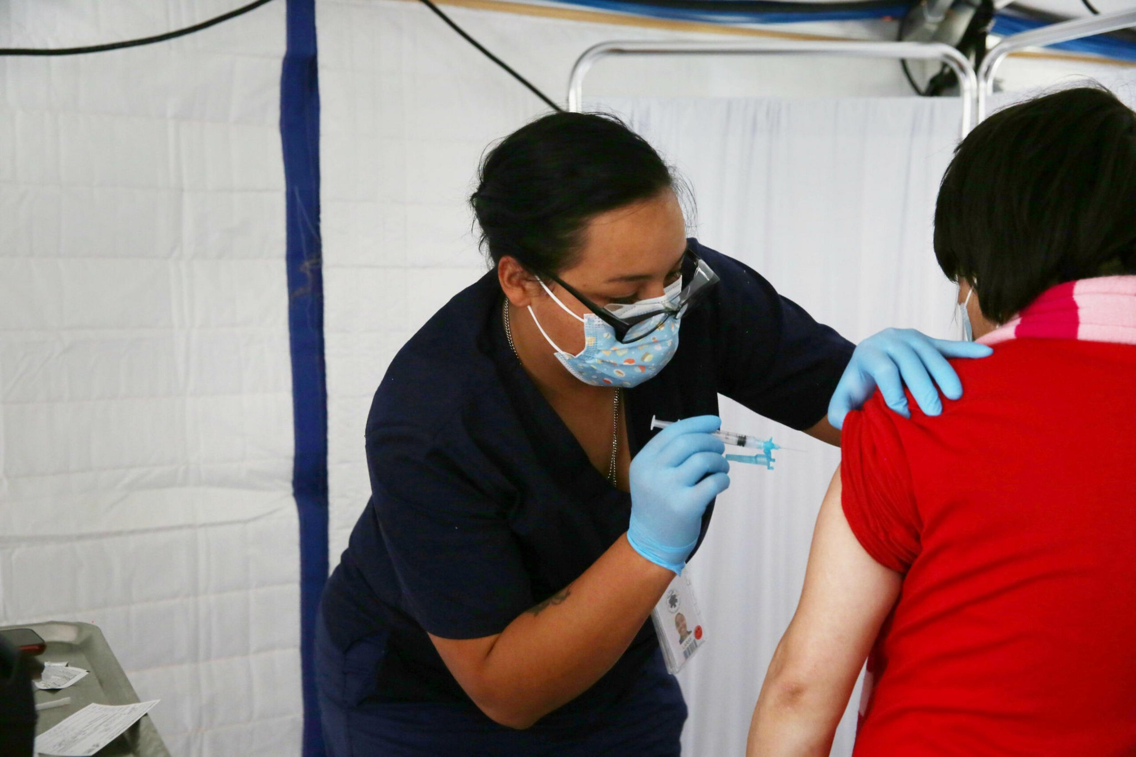 A healthcare worker wearing a mask and gloves administers a vaccine to a person in a red shirt.