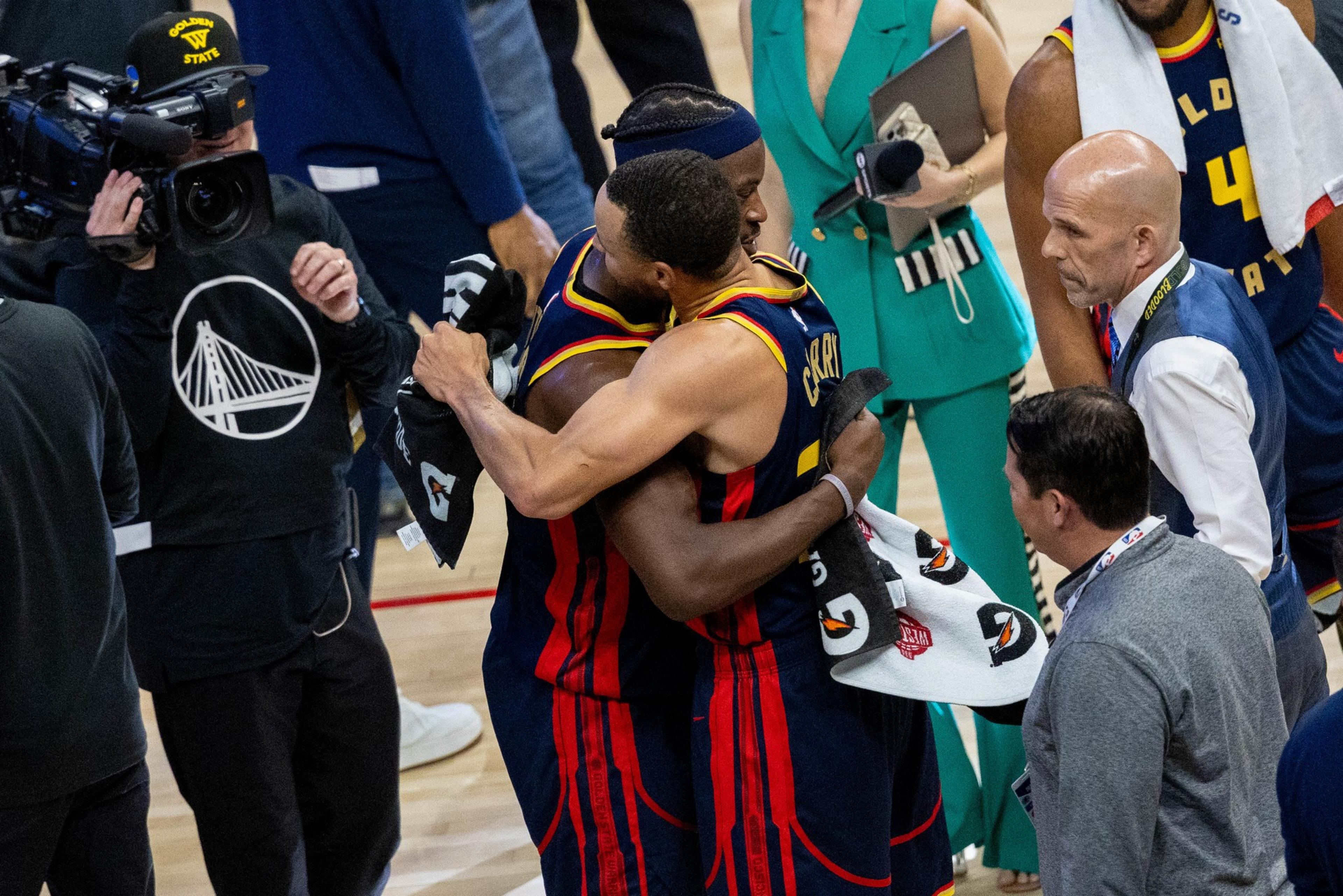 Two basketball players in red and blue jerseys hug surrounded by media and staff. A cameraman captures the moment as a woman in a green outfit stands nearby.