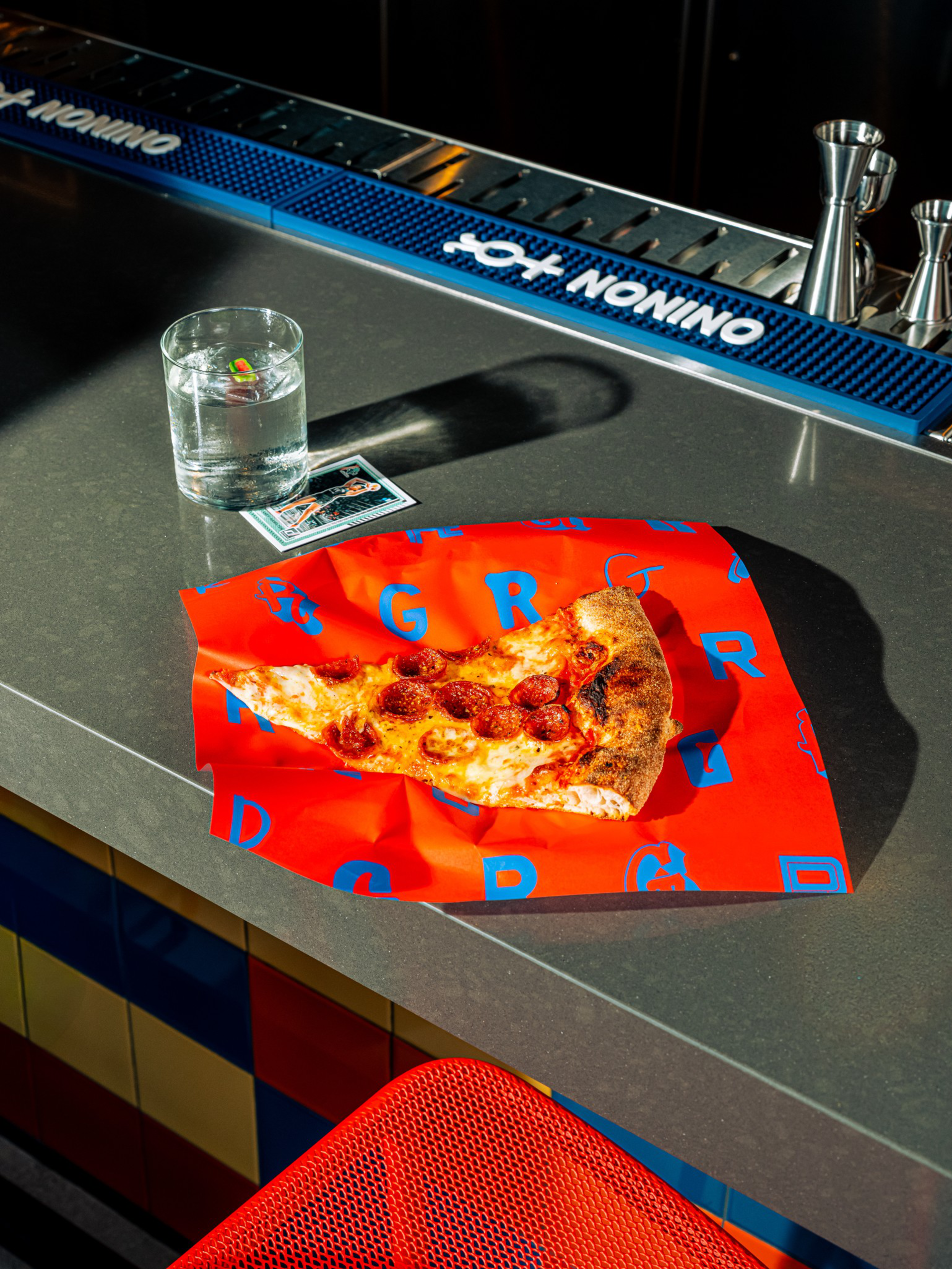 A pepperoni pizza slice rests on bright red paper with blue letters, next to a clear glass of water on a gray bar counter with a red mesh seat nearby.