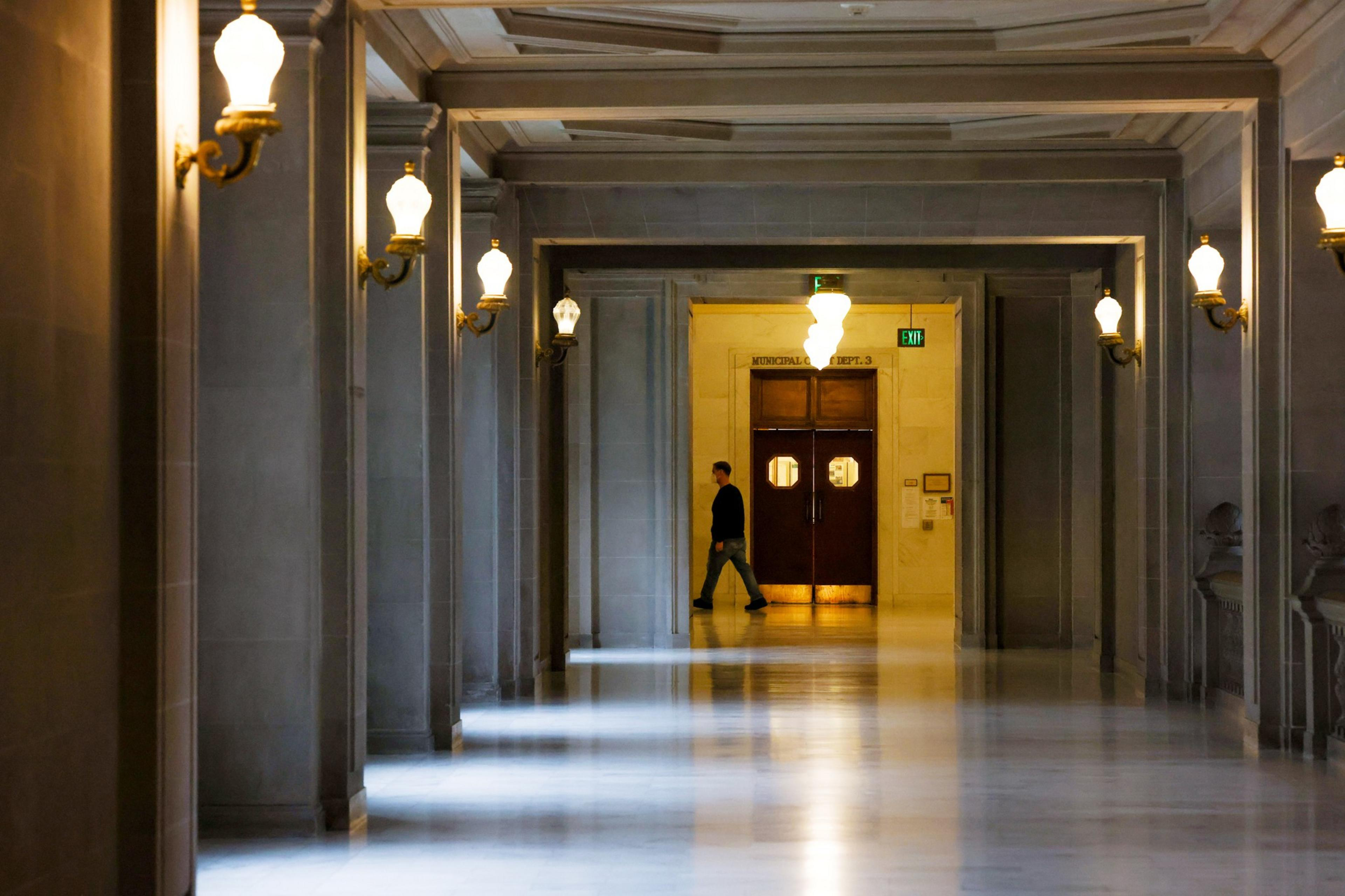 A person walks down a long, dimly lit hallway with ornate wall lamps and a pair of wooden double doors at the far end.