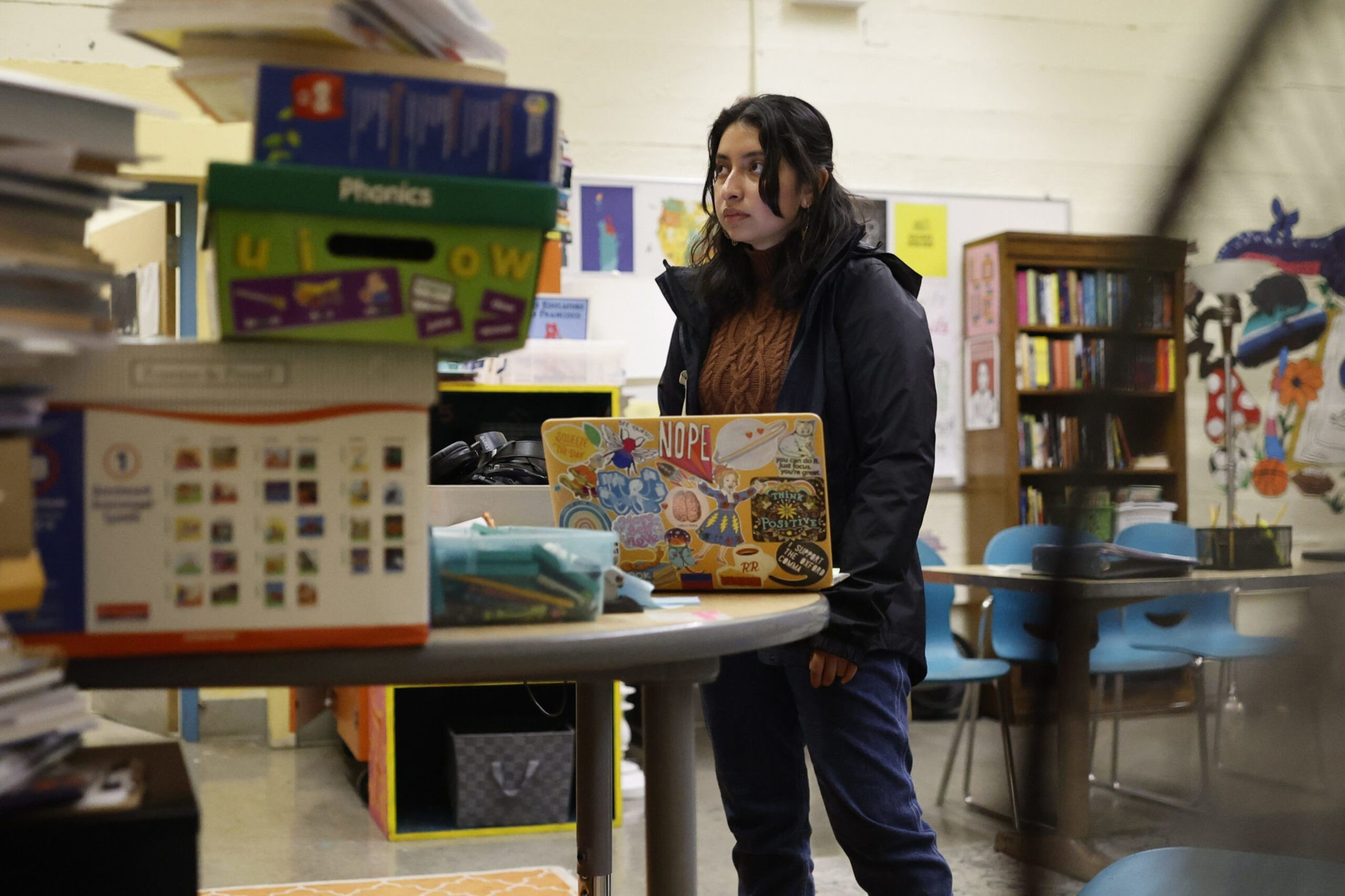 A young woman stands behind a cluttered round table with a colorful laptop in a room filled with books, chairs, and educational materials.