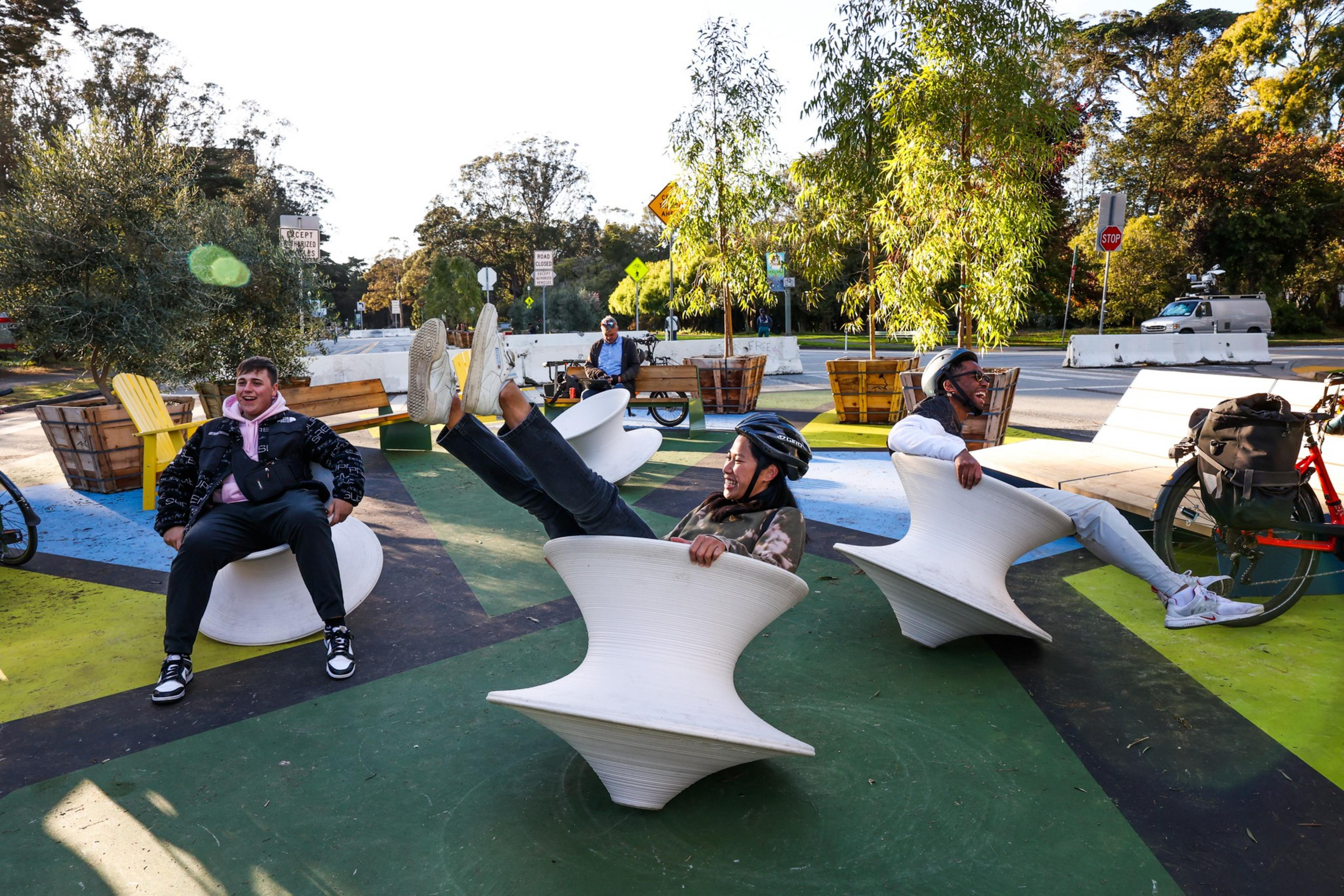 Three people wearing helmets spin happily on white, hourglass-shaped outdoor chairs in a colorful park area with trees, benches, and a bike nearby.