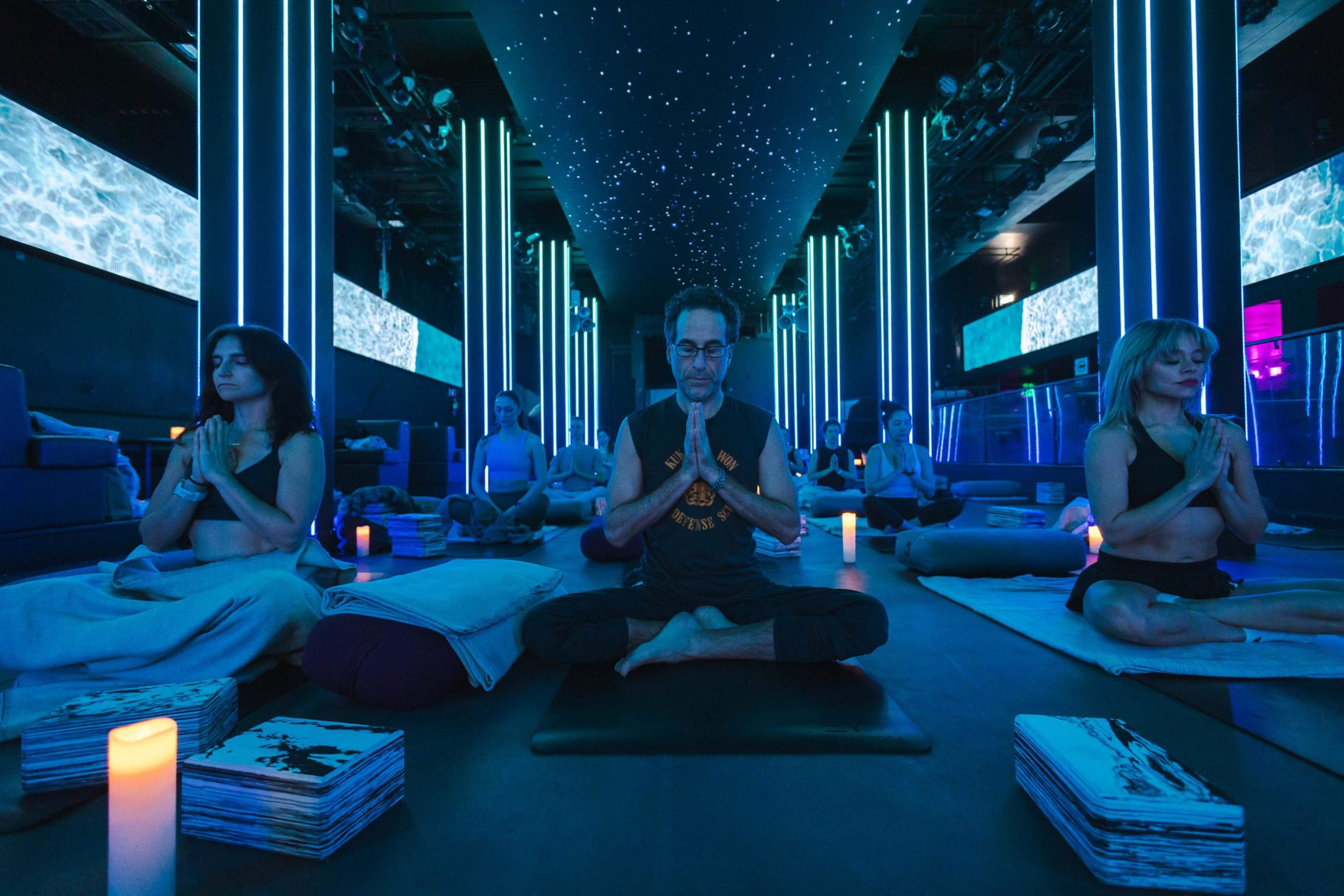 People meditate in a room with blue lighting, LED columns, starry ceiling, and video screens.