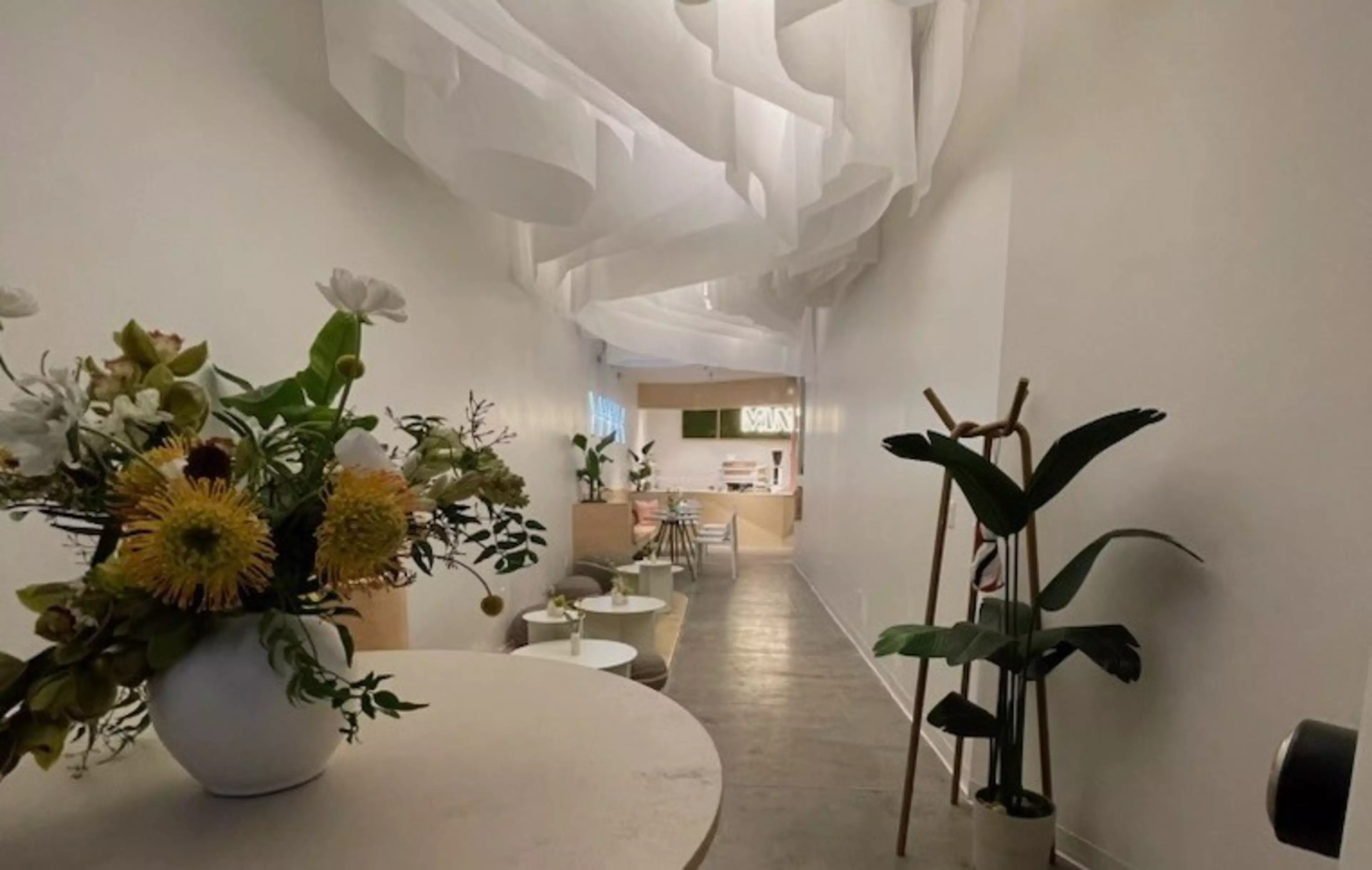 A long, narrow room features white walls, a concrete floor, hanging white fabric above, round tables with flowers, and plants along the right side.