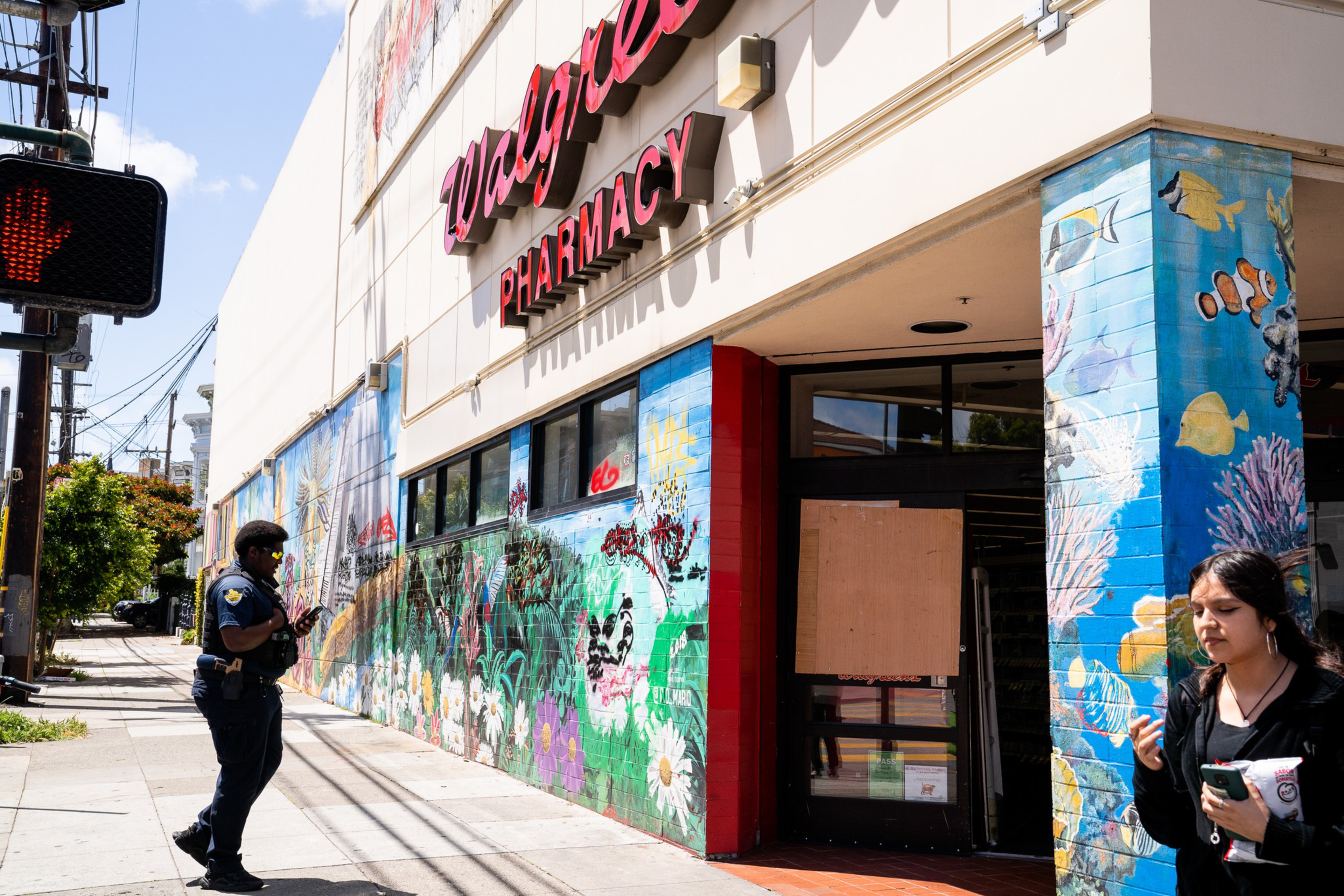 A Walgreens pharmacy with colorful murals on the walls is shown. A police officer is walking by, and a young woman is nearby eating snacks. The entrance has a boarded section.
