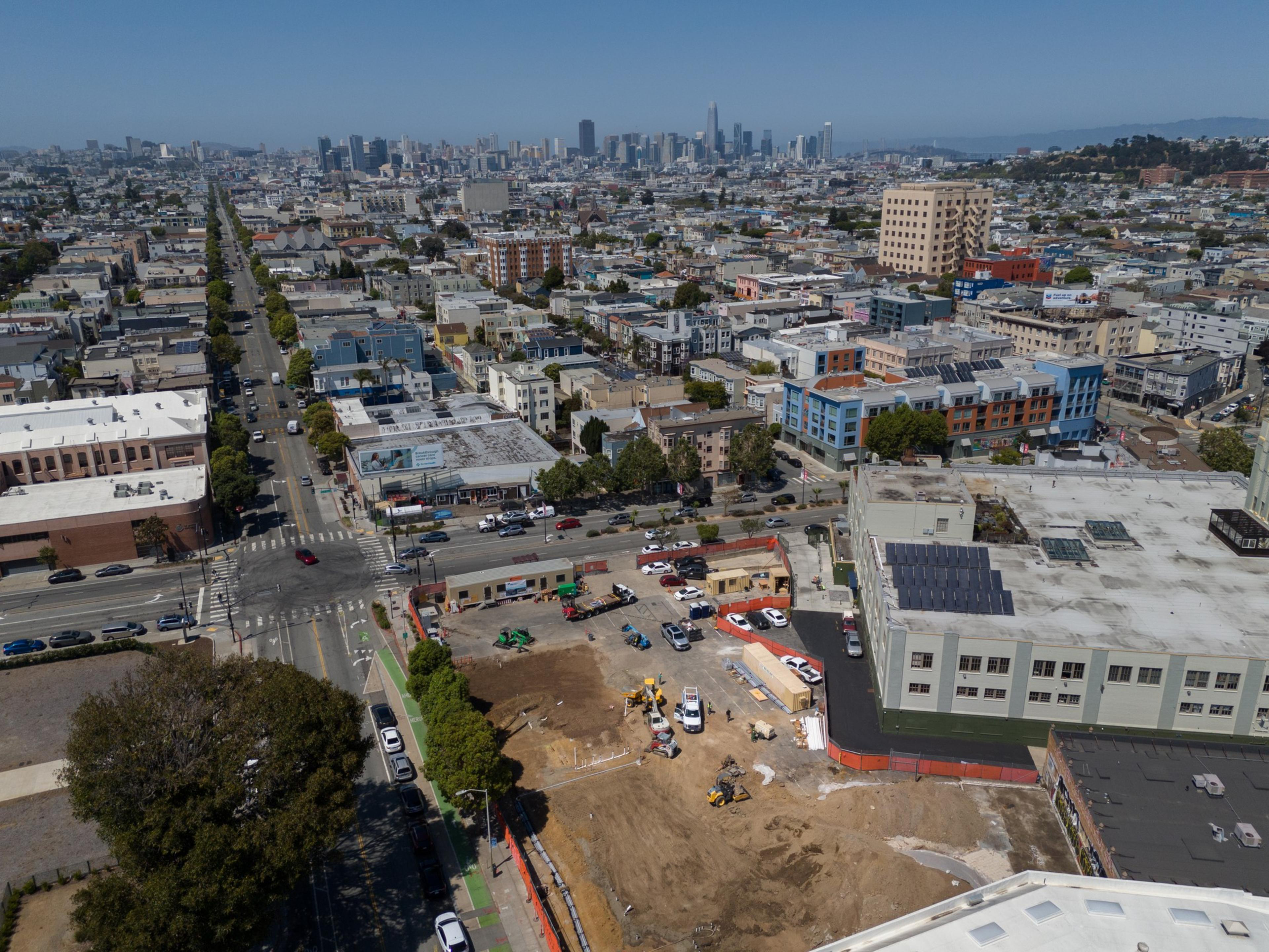 The image shows a cityscape with a construction site in the foreground, intersecting roads, and a grid of buildings extending toward a distant skyline under a clear sky.
