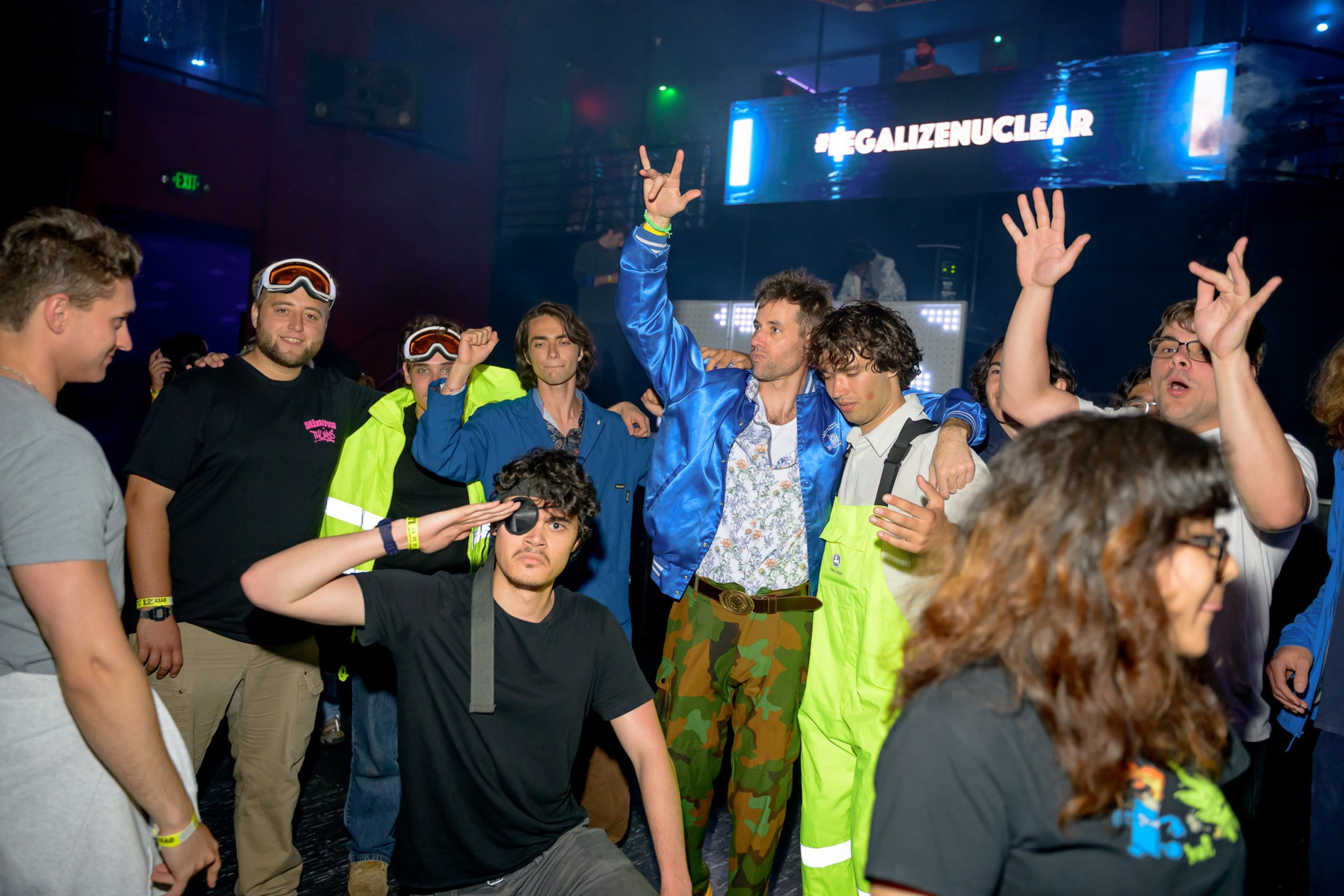 A group of people dressed in brightly colored outfits and costumes are posing energetically at a lively indoor event with colorful lights and a &quot;#LEGALIZENUCLEAR&quot; sign.