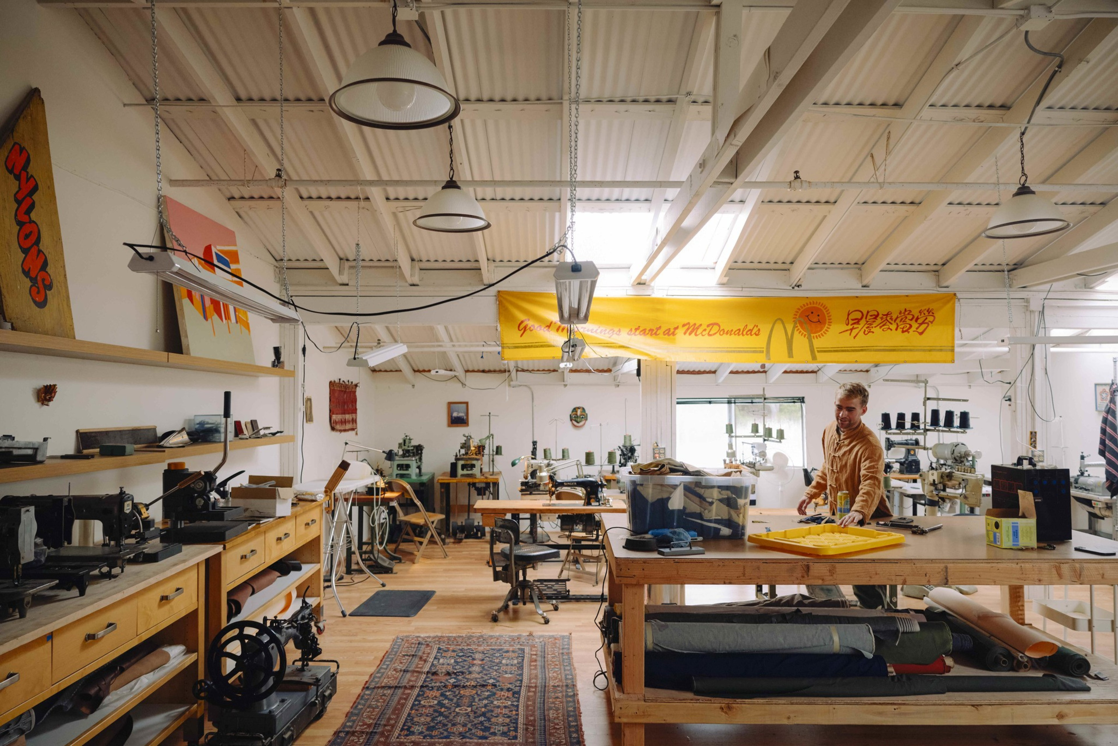 A spacious workshop with sewing machines, fabric rolls, and a person working at a table under bright ceiling lights and a yellow banner.
