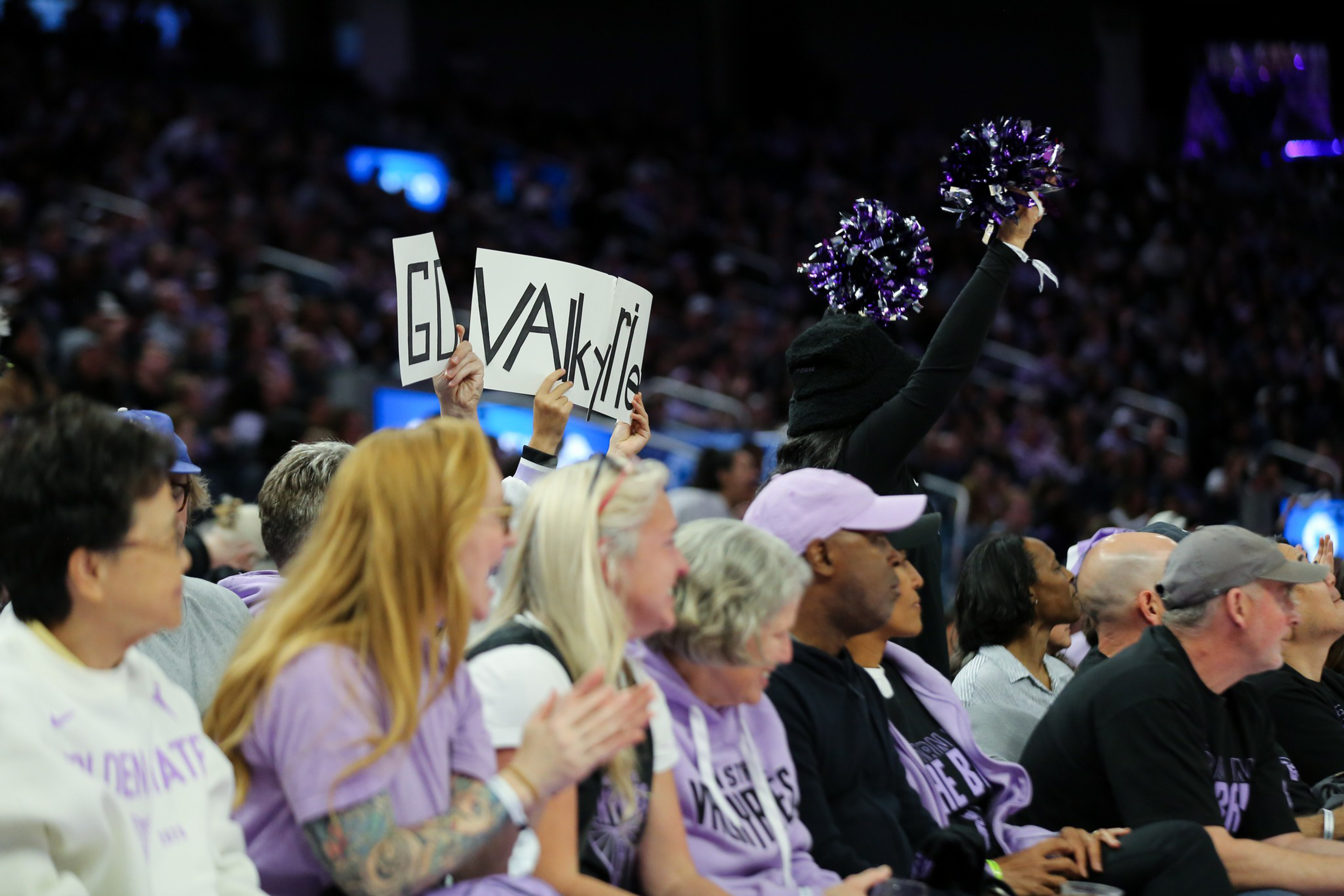 A crowd of spectators watches an event, with one person holding signs and another raising purple pom-poms, all wearing mostly purple and black clothing.
