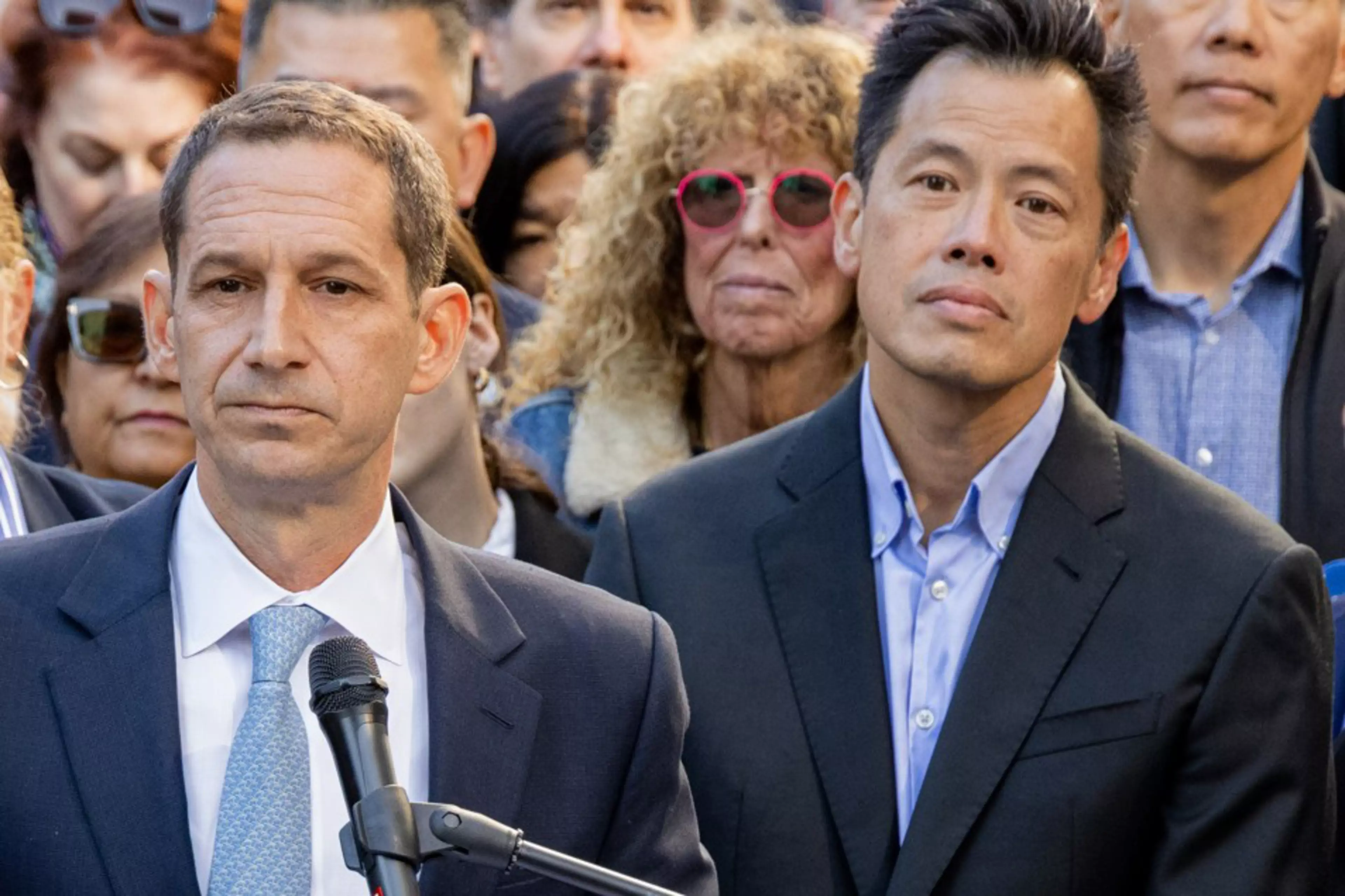 Two men in suits stand solemnly in front of a microphone, with a diverse group of serious-faced people behind them.