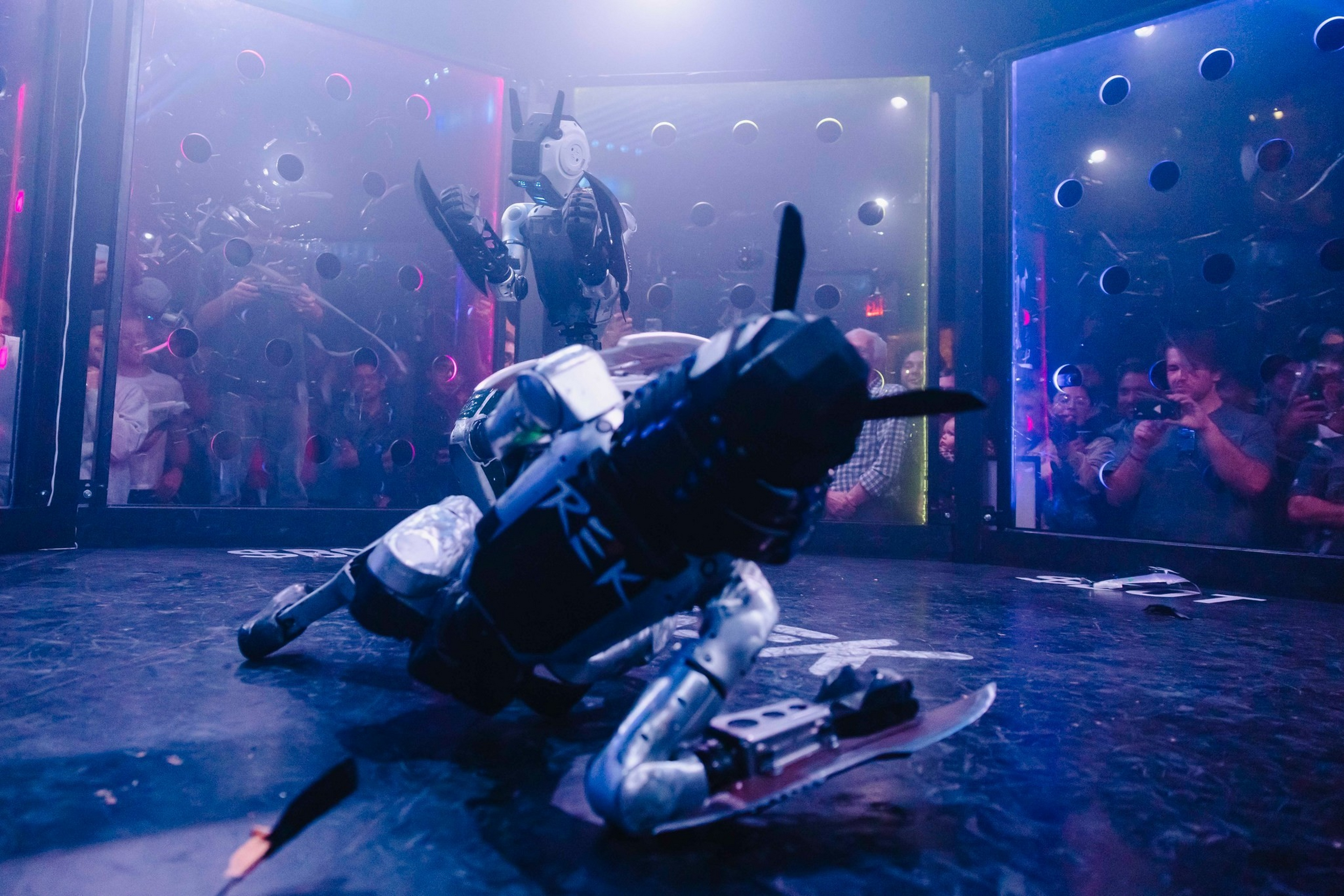 Two humanoid robots are engaged in a fight inside a transparent, circular arena with an audience watching through clear walls.