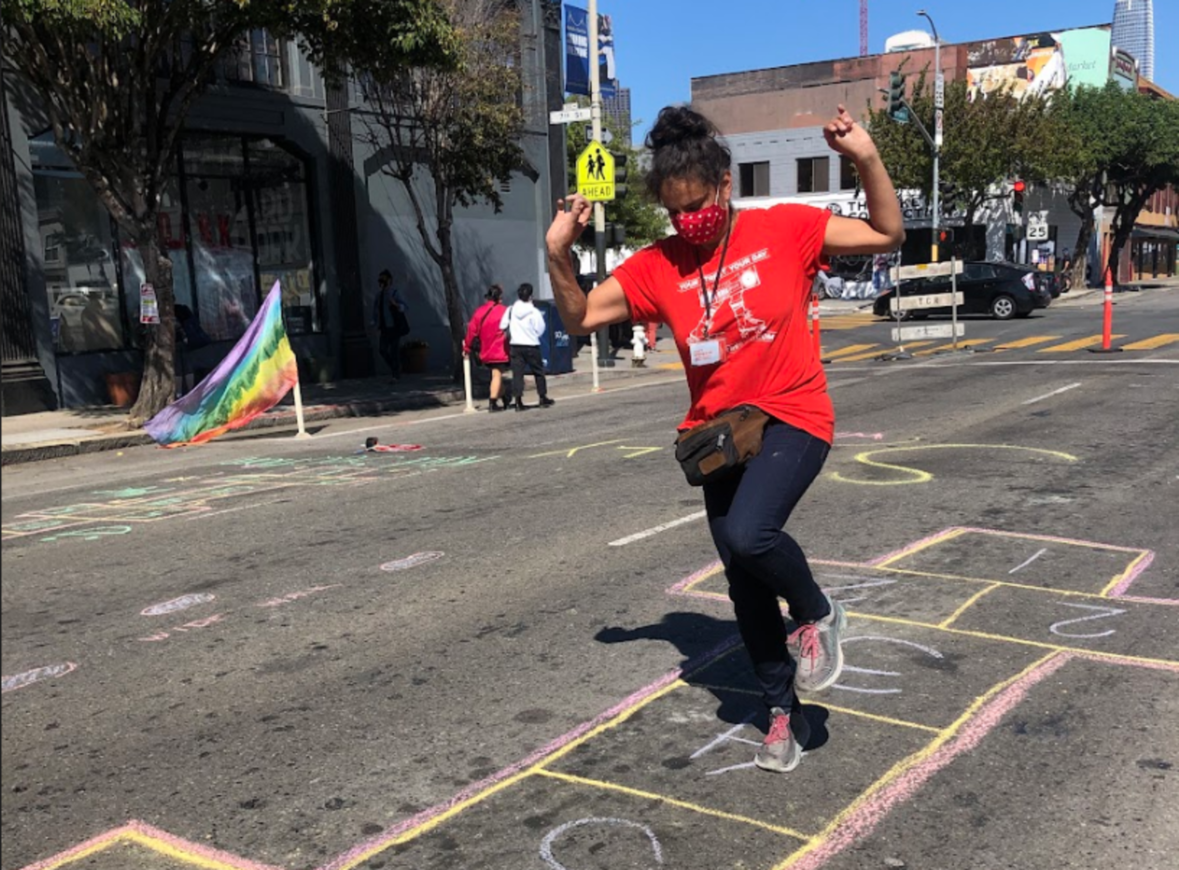 A person wearing a red shirt and mask jumps on a colorful hopscotch drawn with chalk on an urban street on a sunny day.