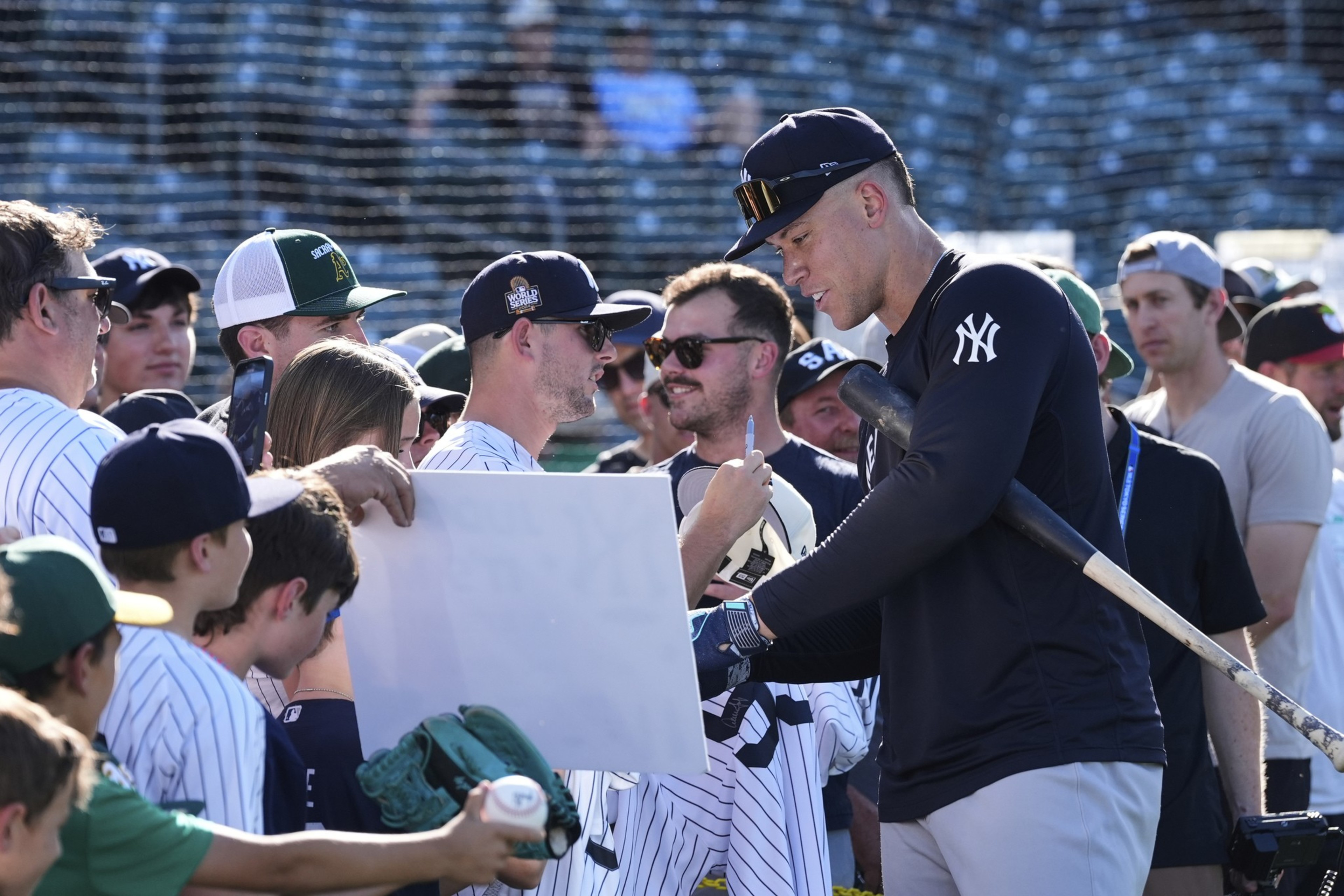 A baseball player wearing a Yankees uniform signs autographs for fans, including a child with a green glove and baseball, in a stadium setting.
