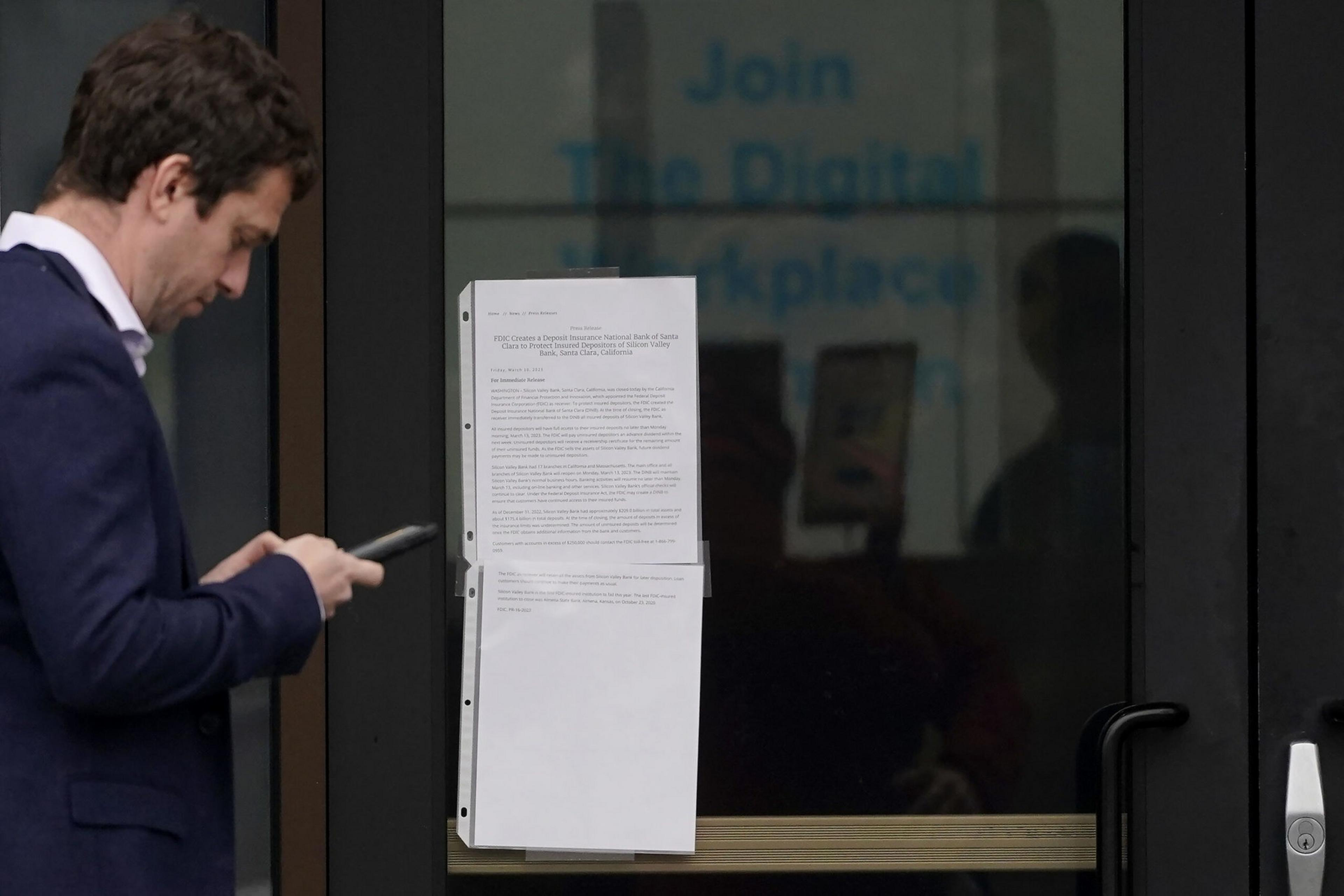 A man in a blue suit looks at his phone next to a glass door with printed papers taped to it and a faint "Join The Digital Workplace" sign inside.