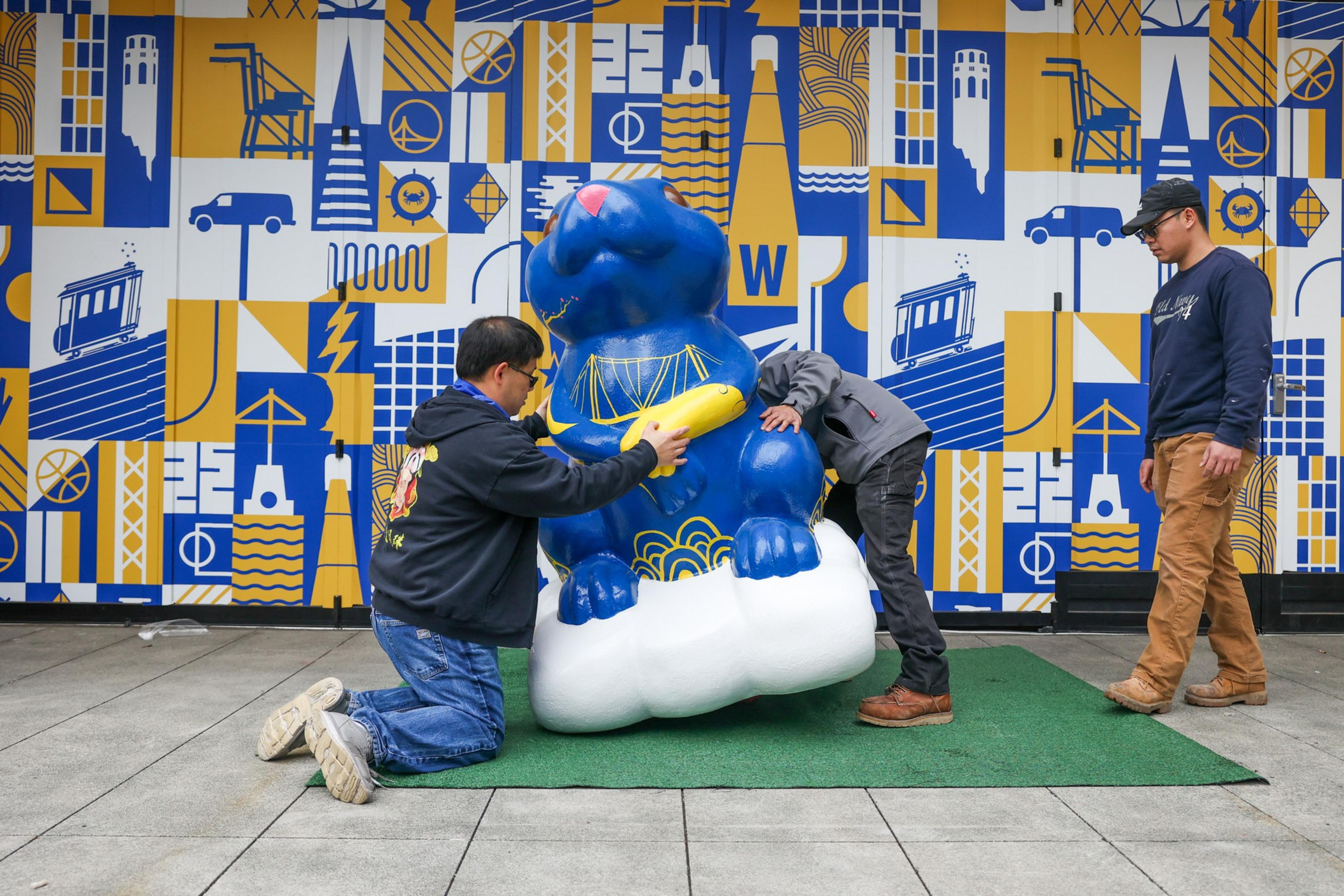 A blue statue of a dog holding a yellow object sits on a white cloud base as three men interact with it against a blue and yellow graphic wall.