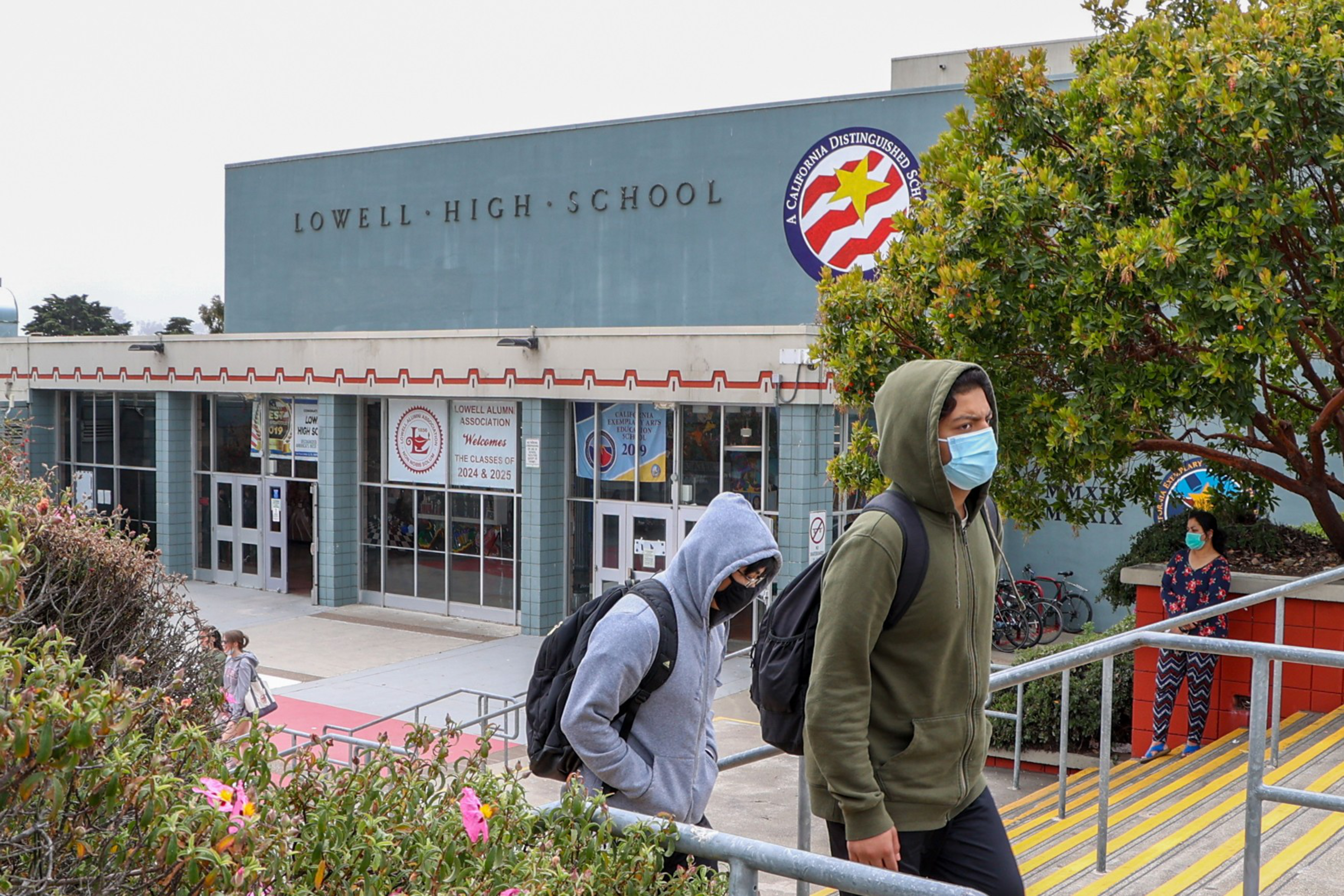 Students wearing masks and hoodies walk past Lowell High School on a cloudy day, with some greenery and a staff member visible near the entrance.