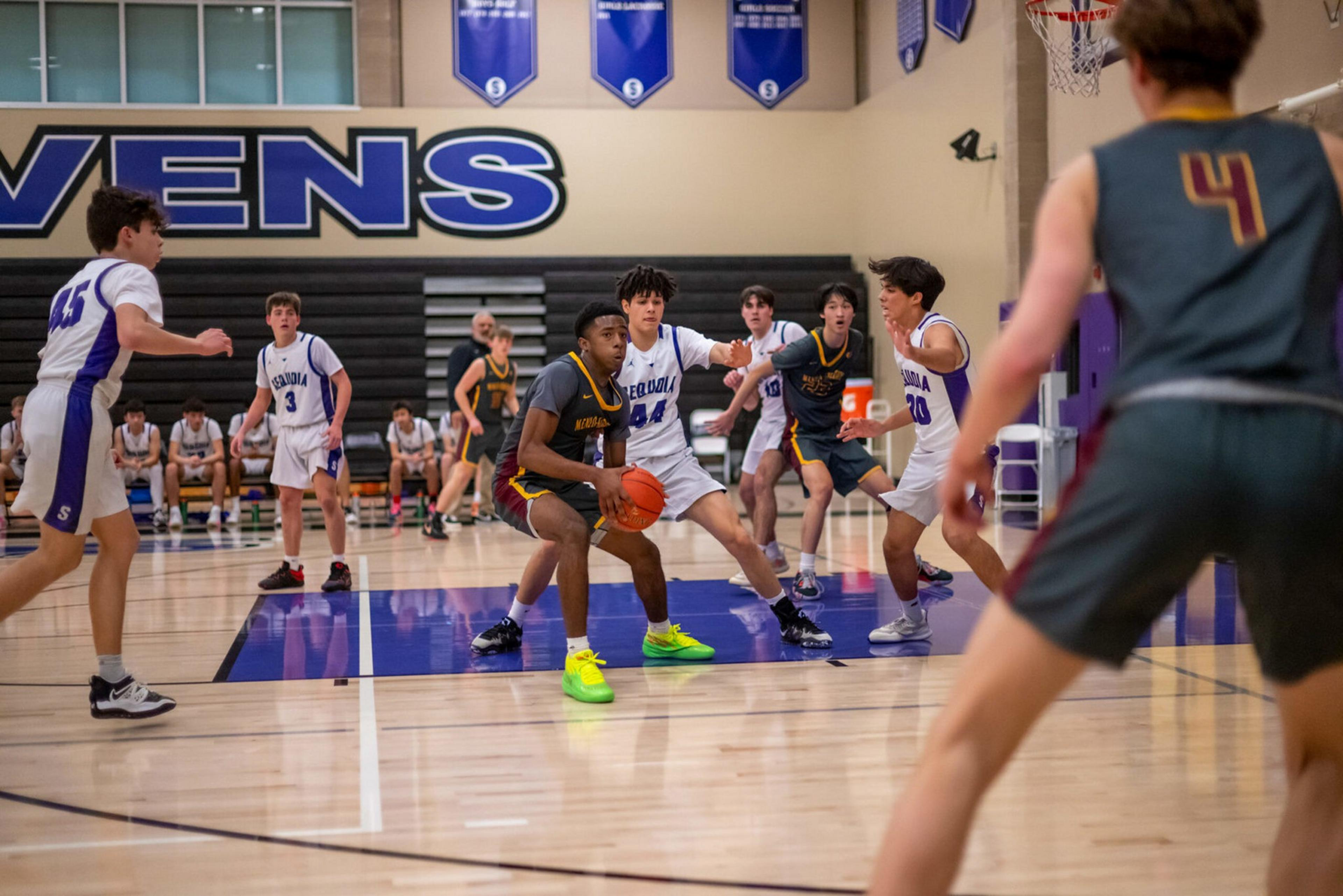 A basketball player in dark uniform holds the ball while being closely guarded by three players in white uniforms near the basket on a gym court.