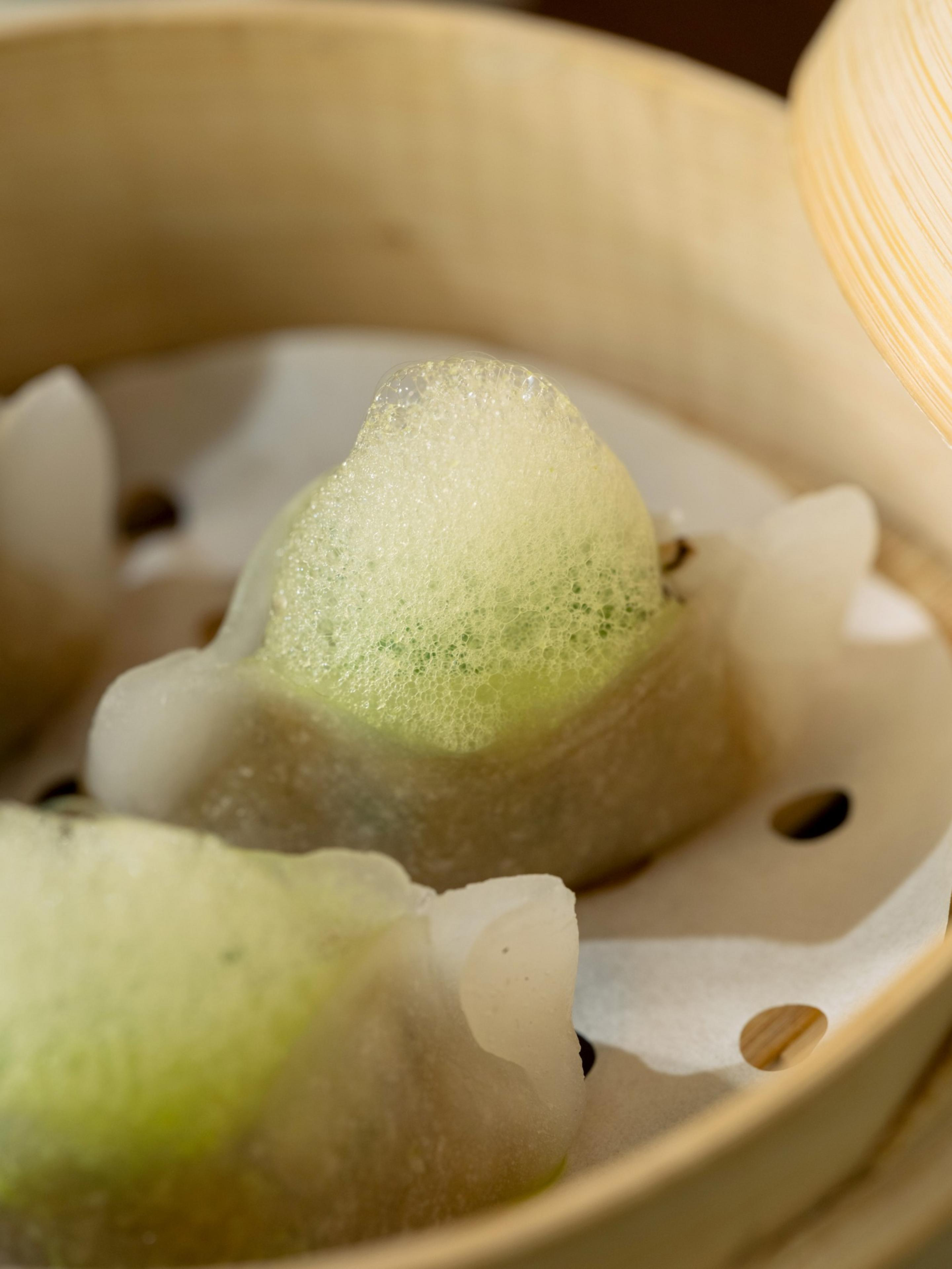 Translucent dumplings with green foam on top rest on parchment paper inside a bamboo steamer basket.