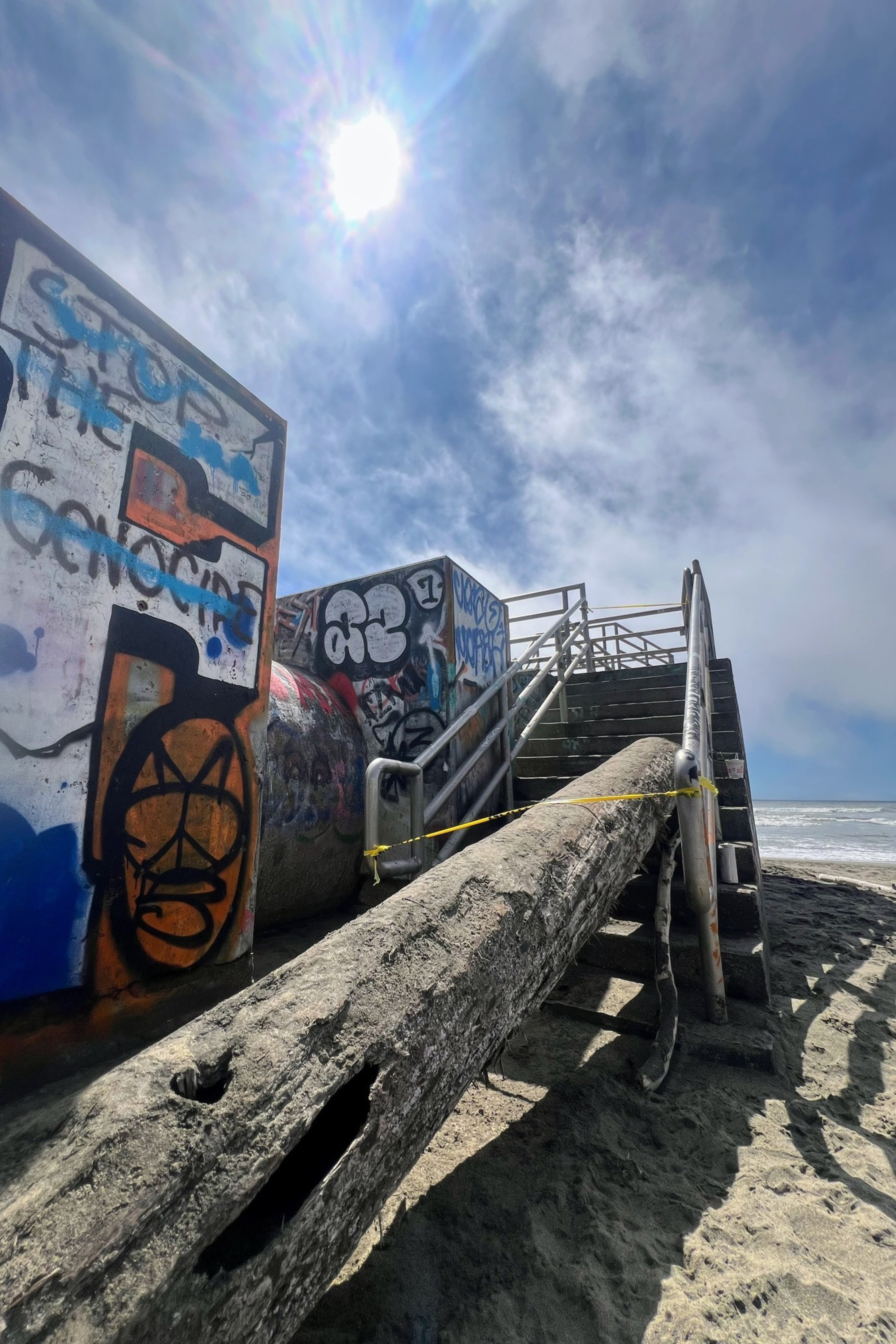 The image shows a sandy beach with graffiti-covered structures and a staircase blocked by a large log, under a bright sun and a partly cloudy sky.