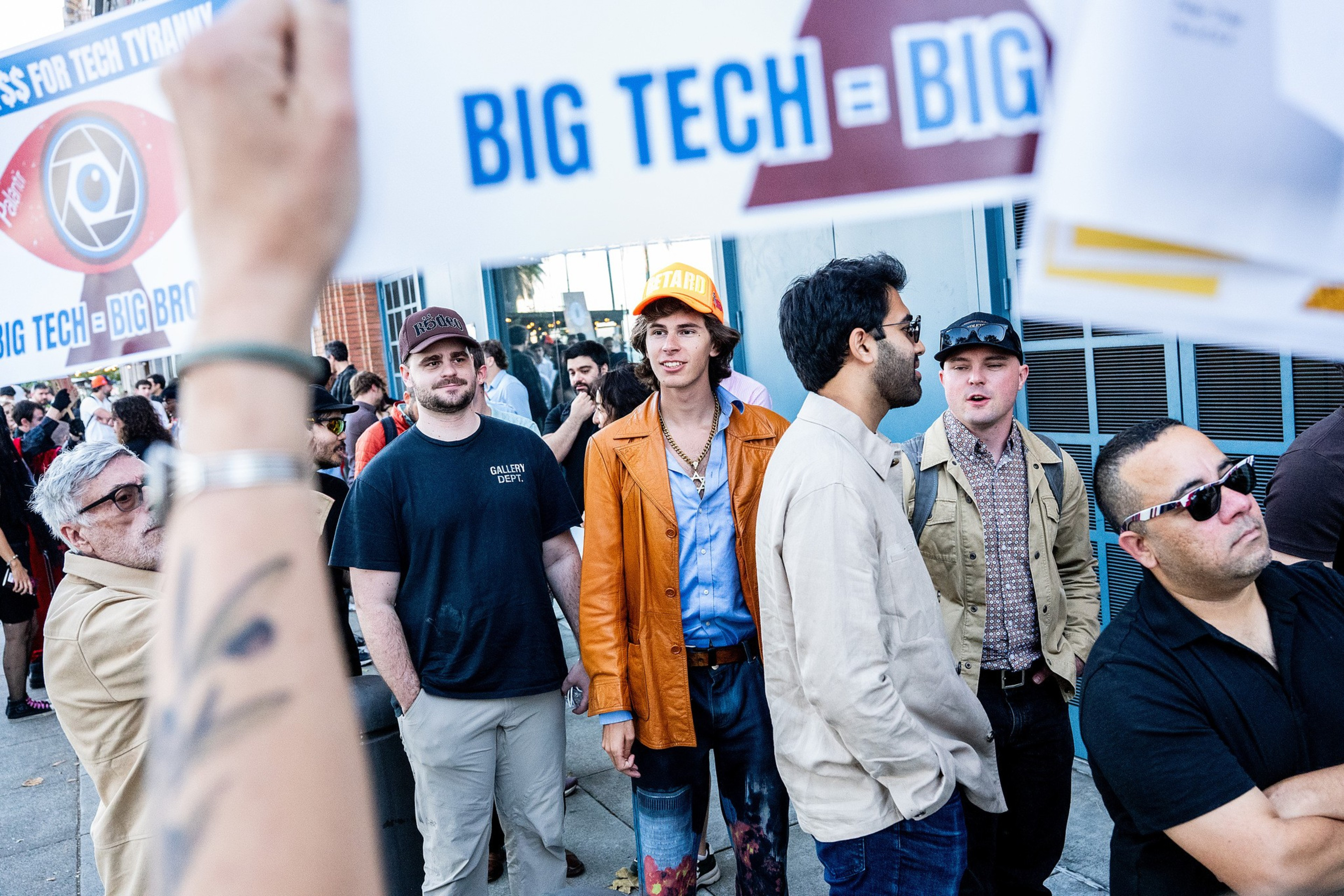 A group of people stands outside, some holding signs with messages against big tech companies. One person wears an orange jacket and hat.