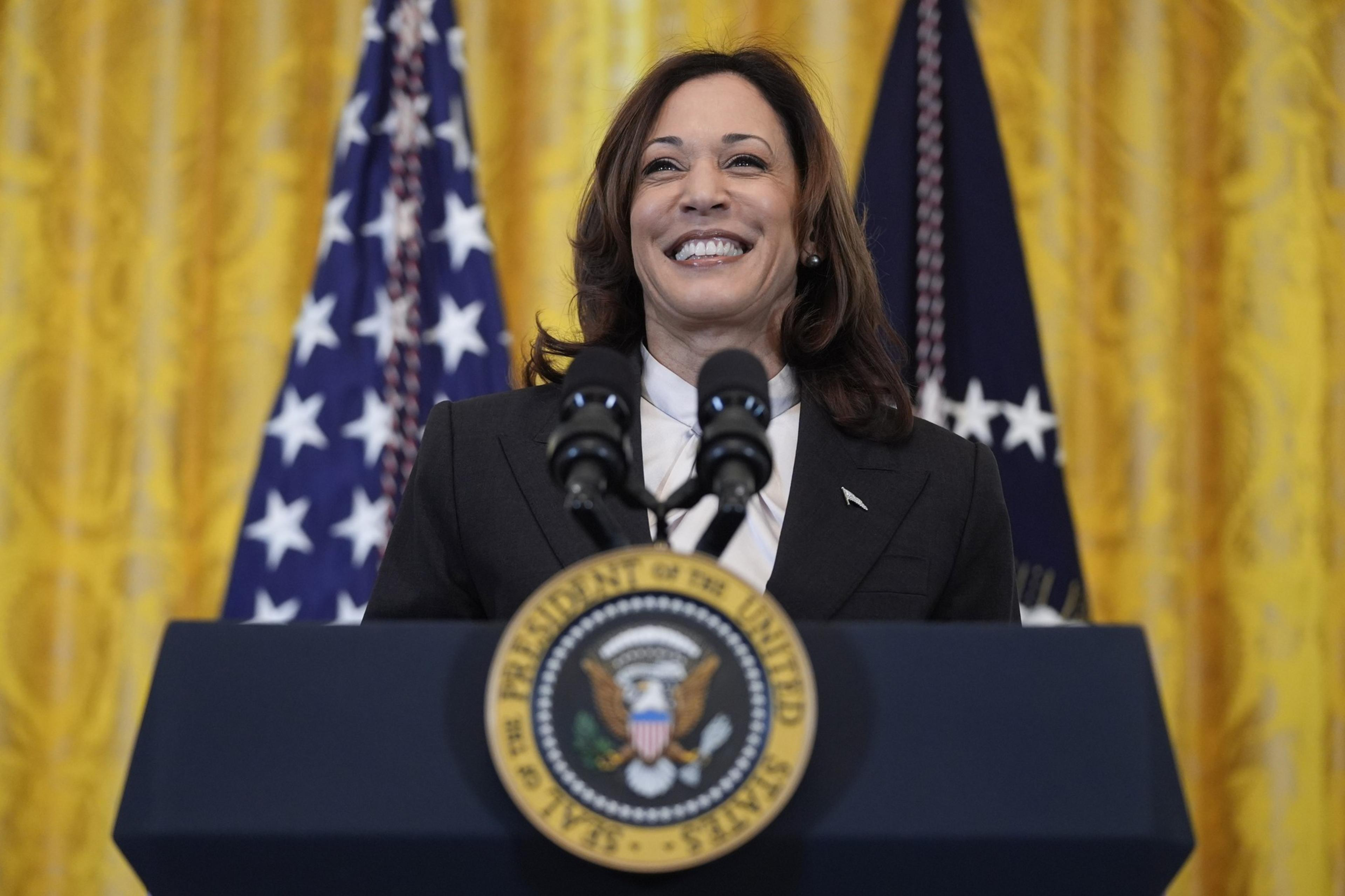 A woman is smiling while standing at a podium with the U.S. presidential seal. Behind her are two U.S. flags and yellow curtains.