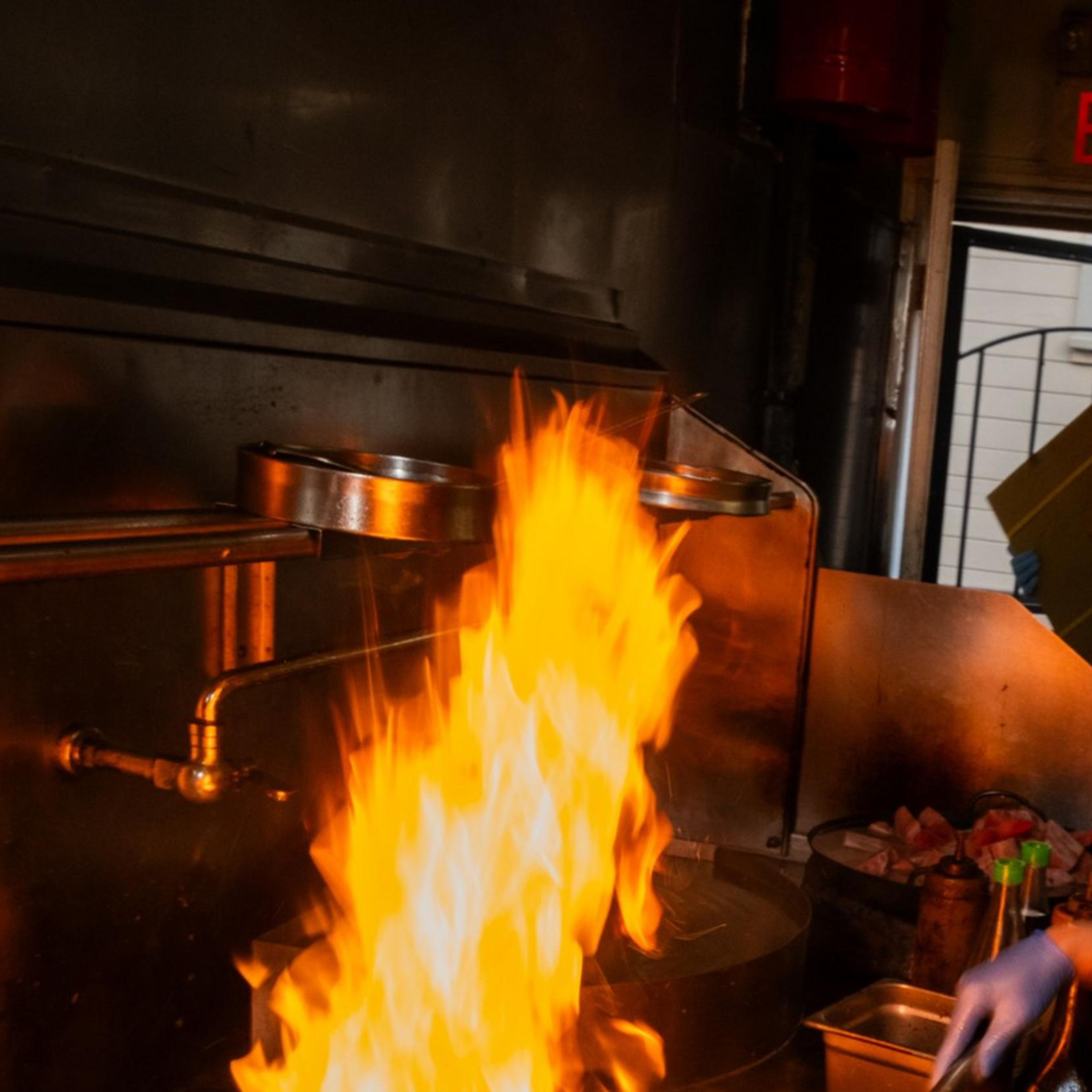 A person in a red bandana is cooking in a kitchen, smiling while stirring food in a flaming pan. The kitchen has shelves with ingredients and an "EXIT" sign visible.