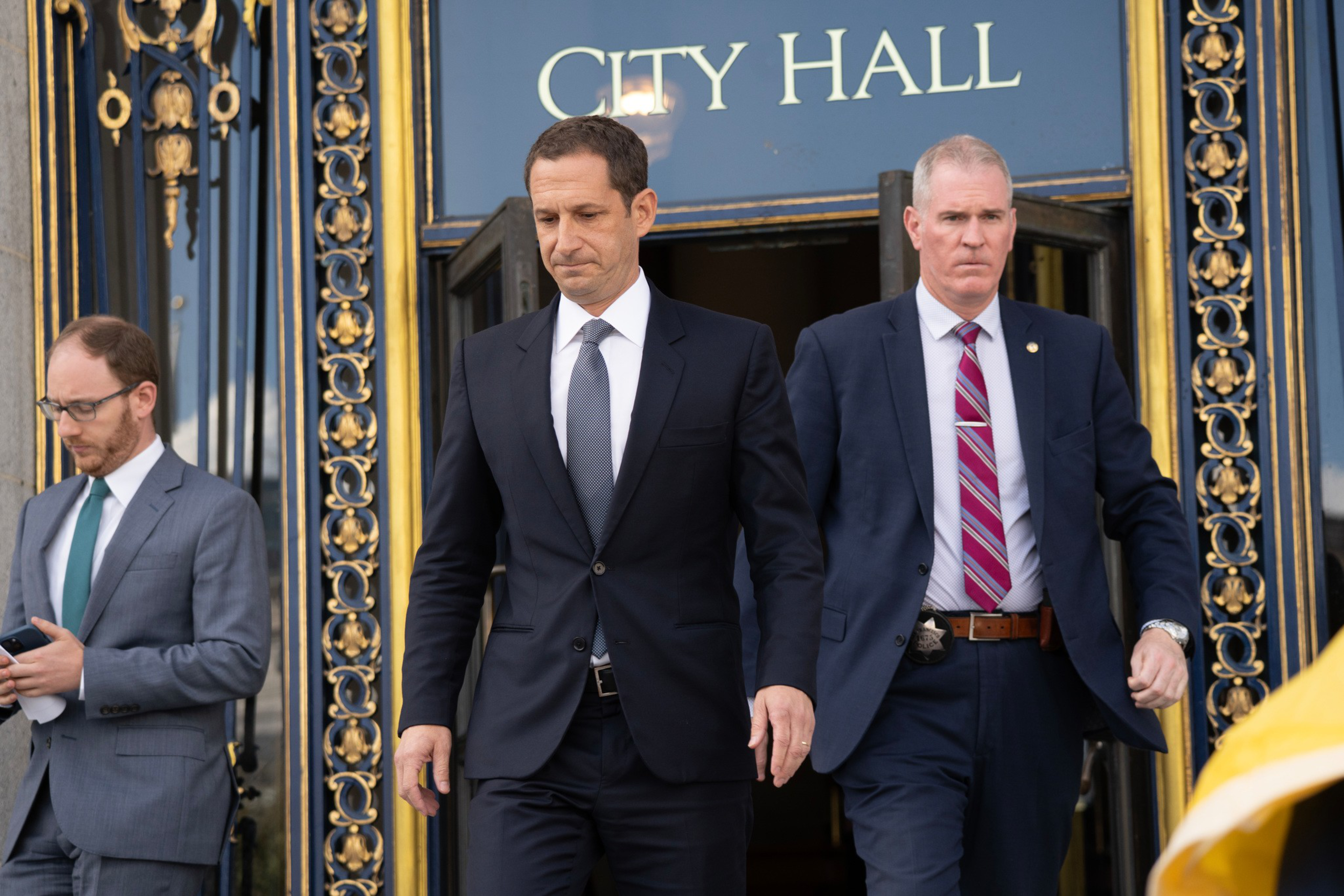 Three men in suits walk down ornate steps outside a City Hall building, with two men looking serious and one focused on his phone.