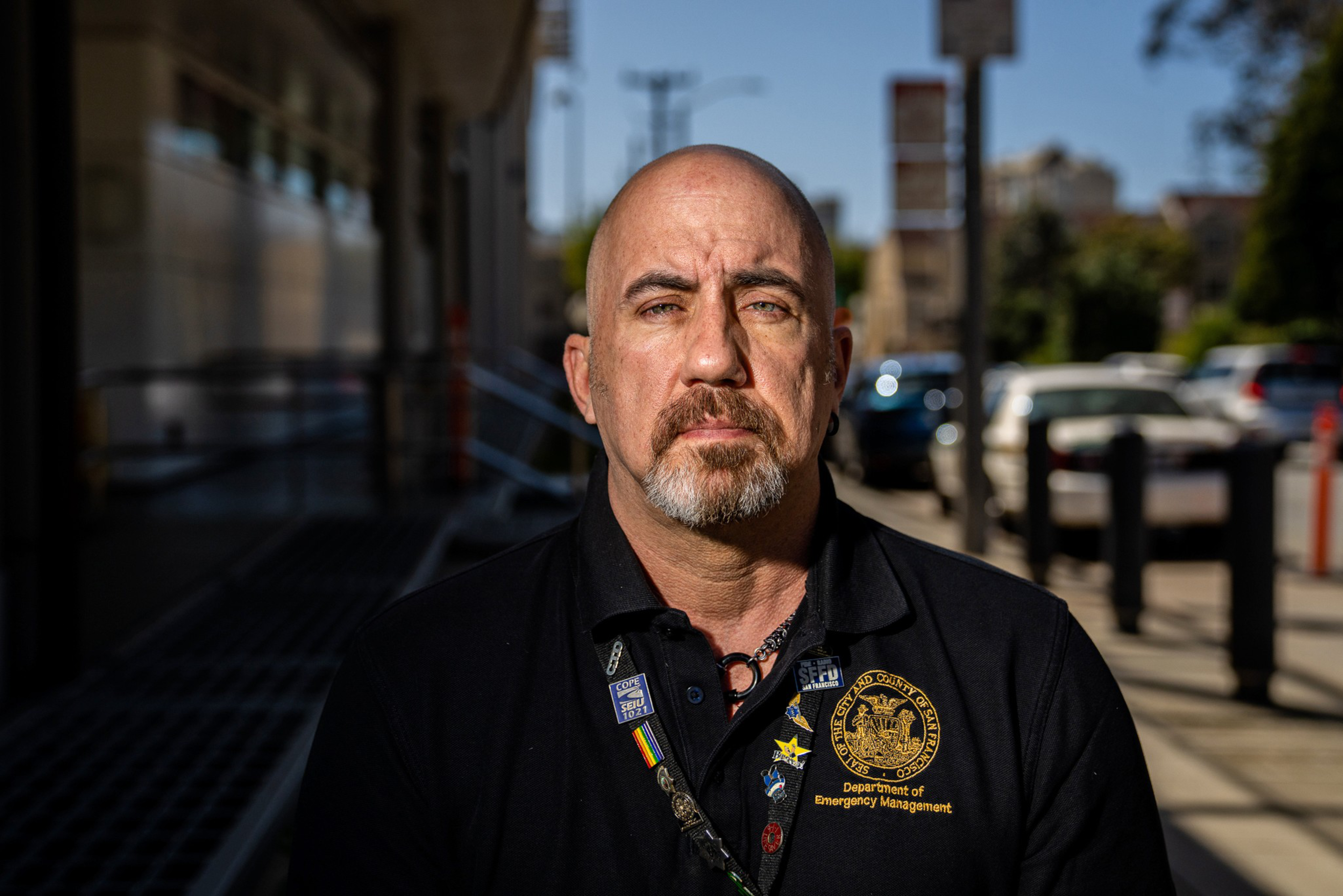 A serious-looking bald man with a goatee wears a black Department of Emergency Management shirt and stands outside on a sunny day.