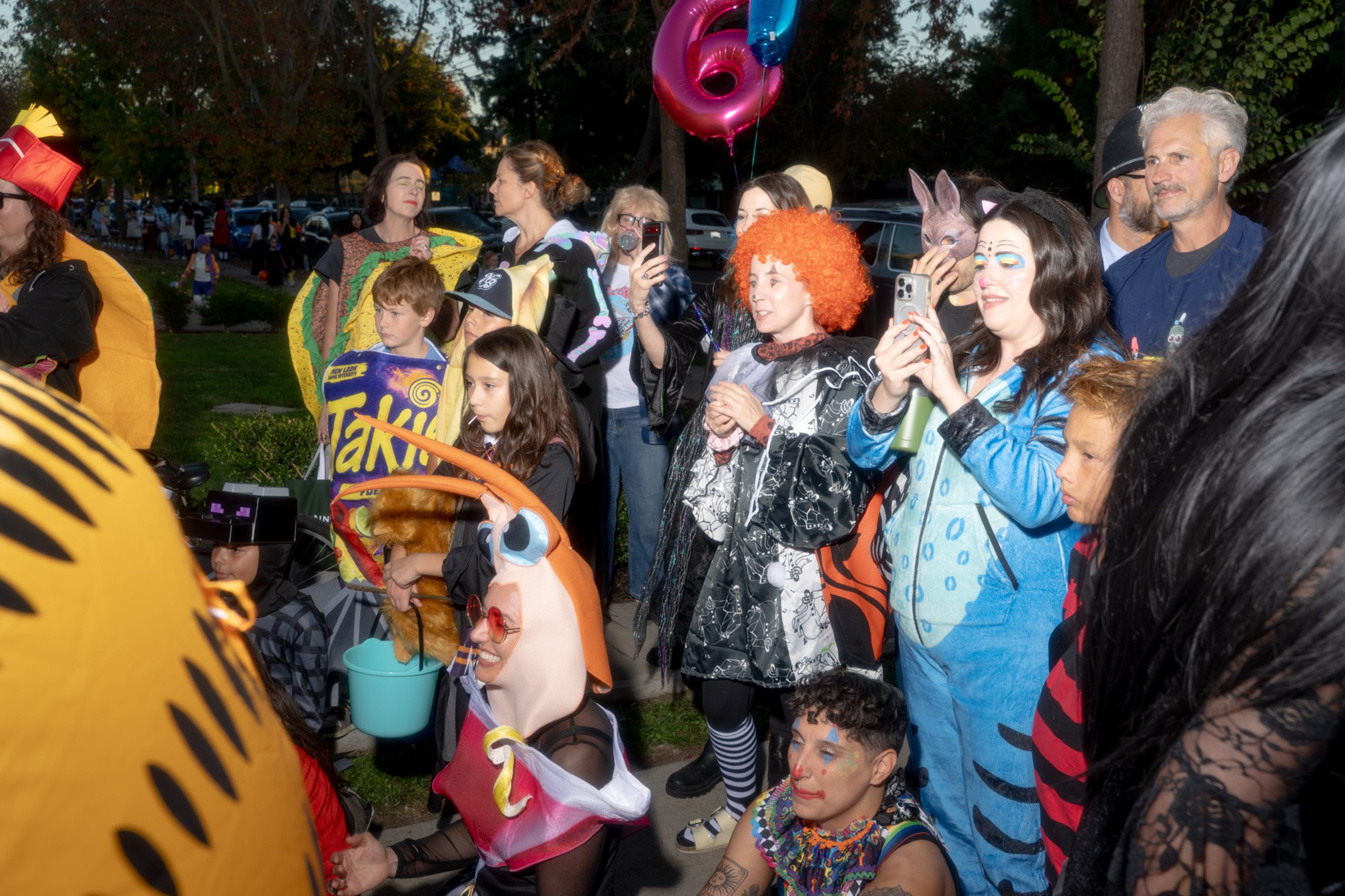 A group of children and adults in colorful costumes, including a Takis chip bag, blue onesie, clown wig, and shrimp outfit, gather outdoors at dusk.