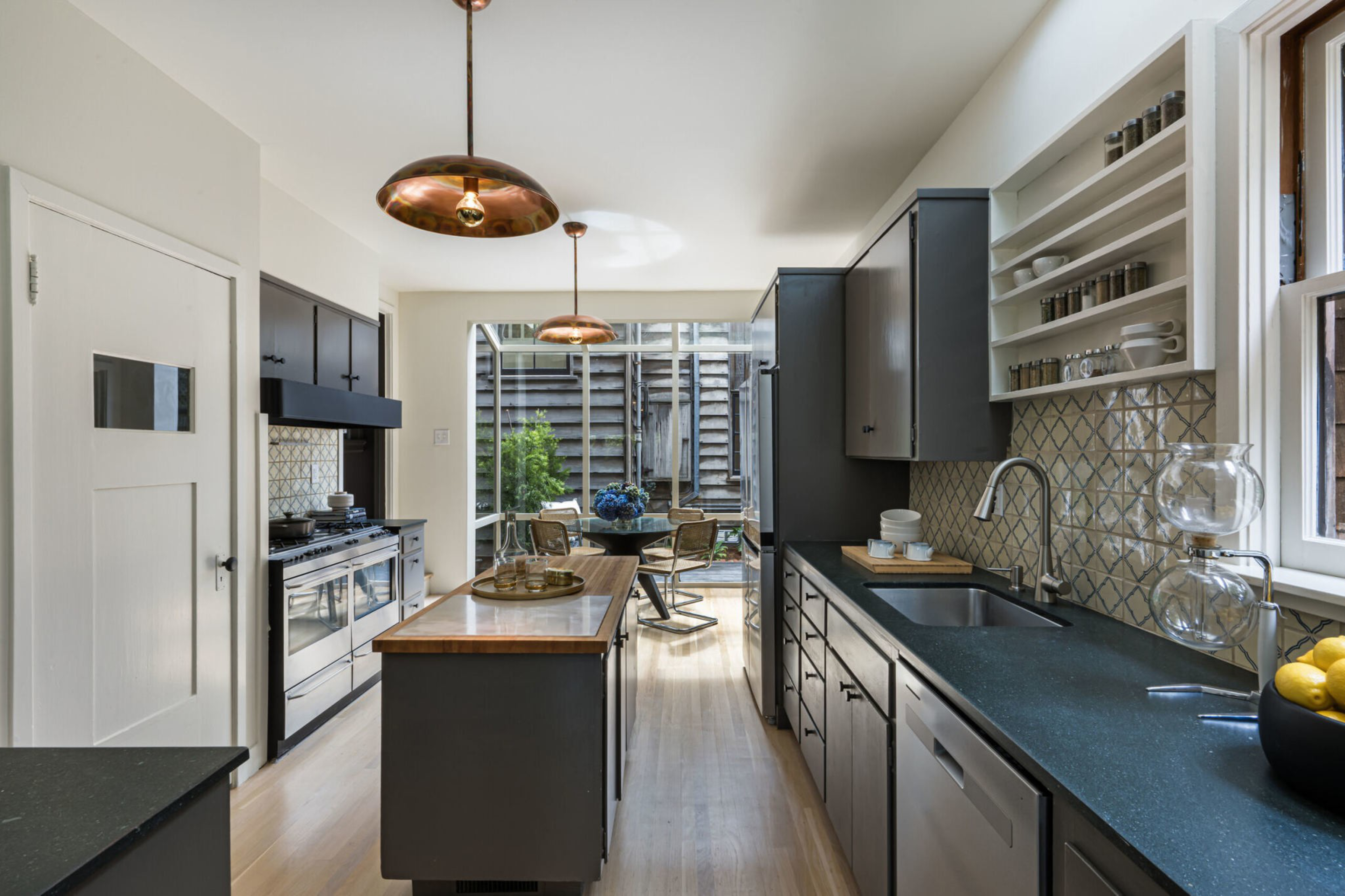 A sleek kitchen features dark gray cabinets, a central wooden island, black countertops, copper pendant lights, and a dining area with glass walls in the background.