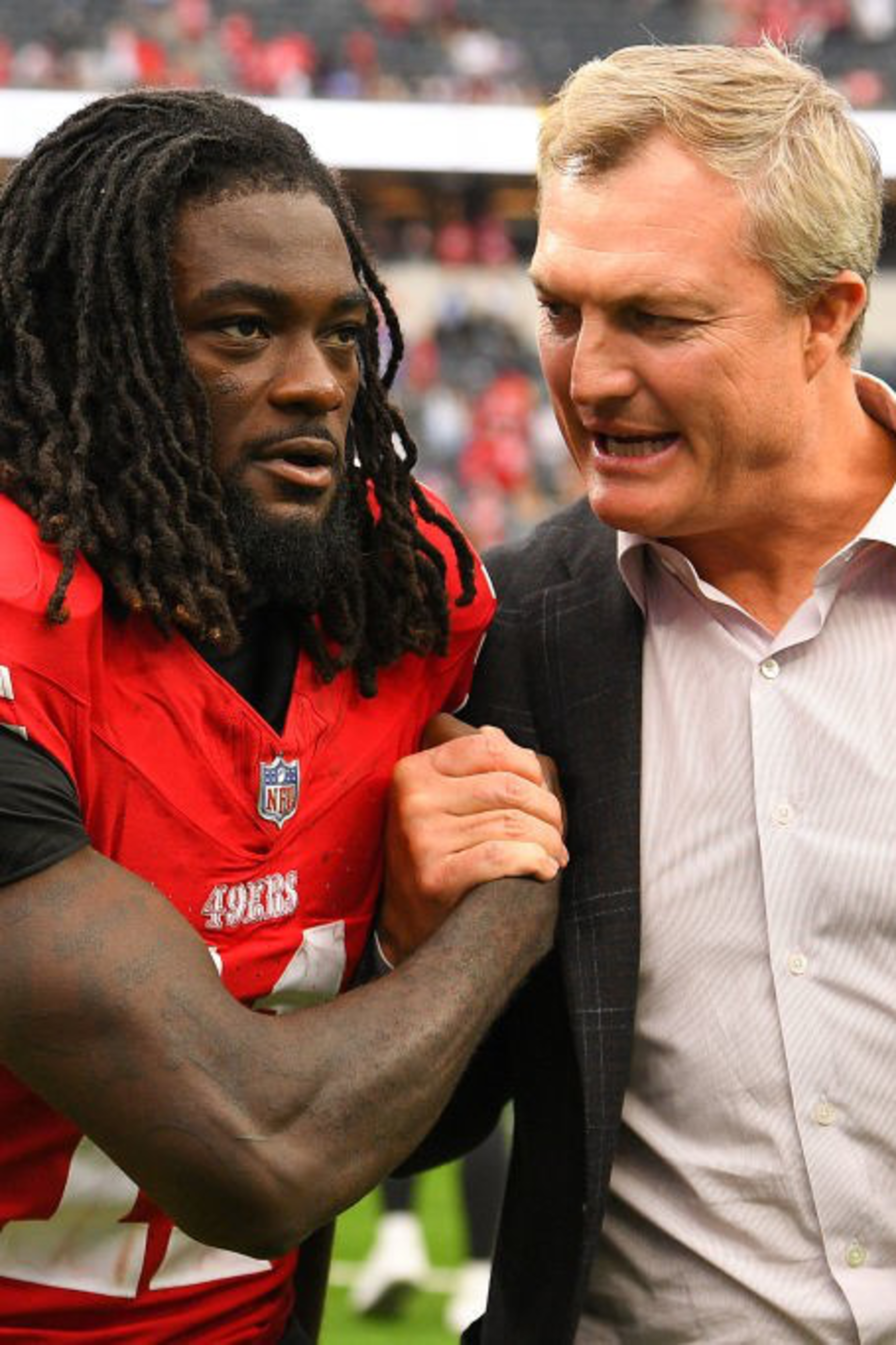 A football player in a red jersey shakes hands with a man in a suit on a field, with blurred spectators in the background.