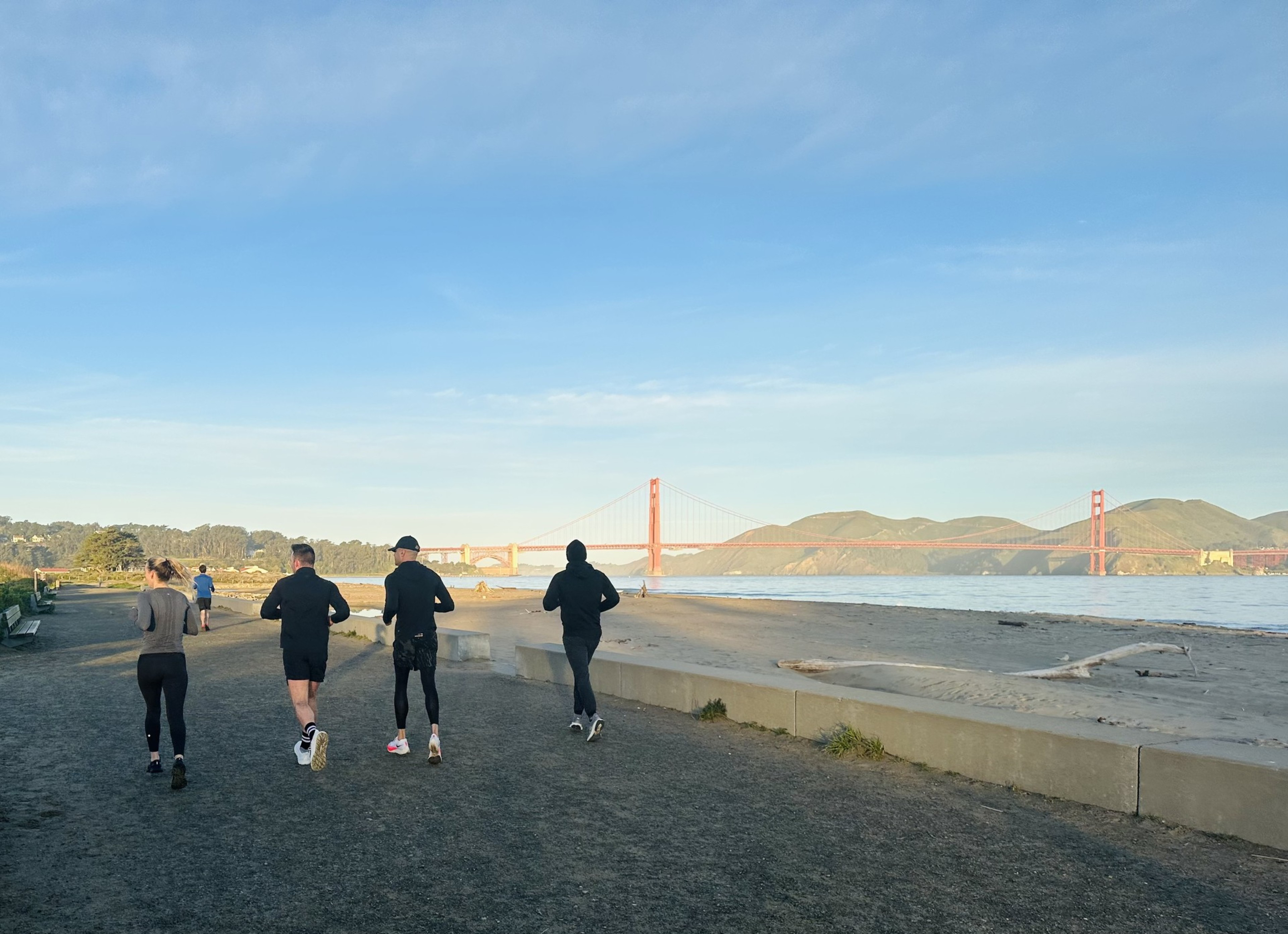 Four people jog along a beachside path on a sunny day with the iconic Golden Gate Bridge and rolling hills in the background.
