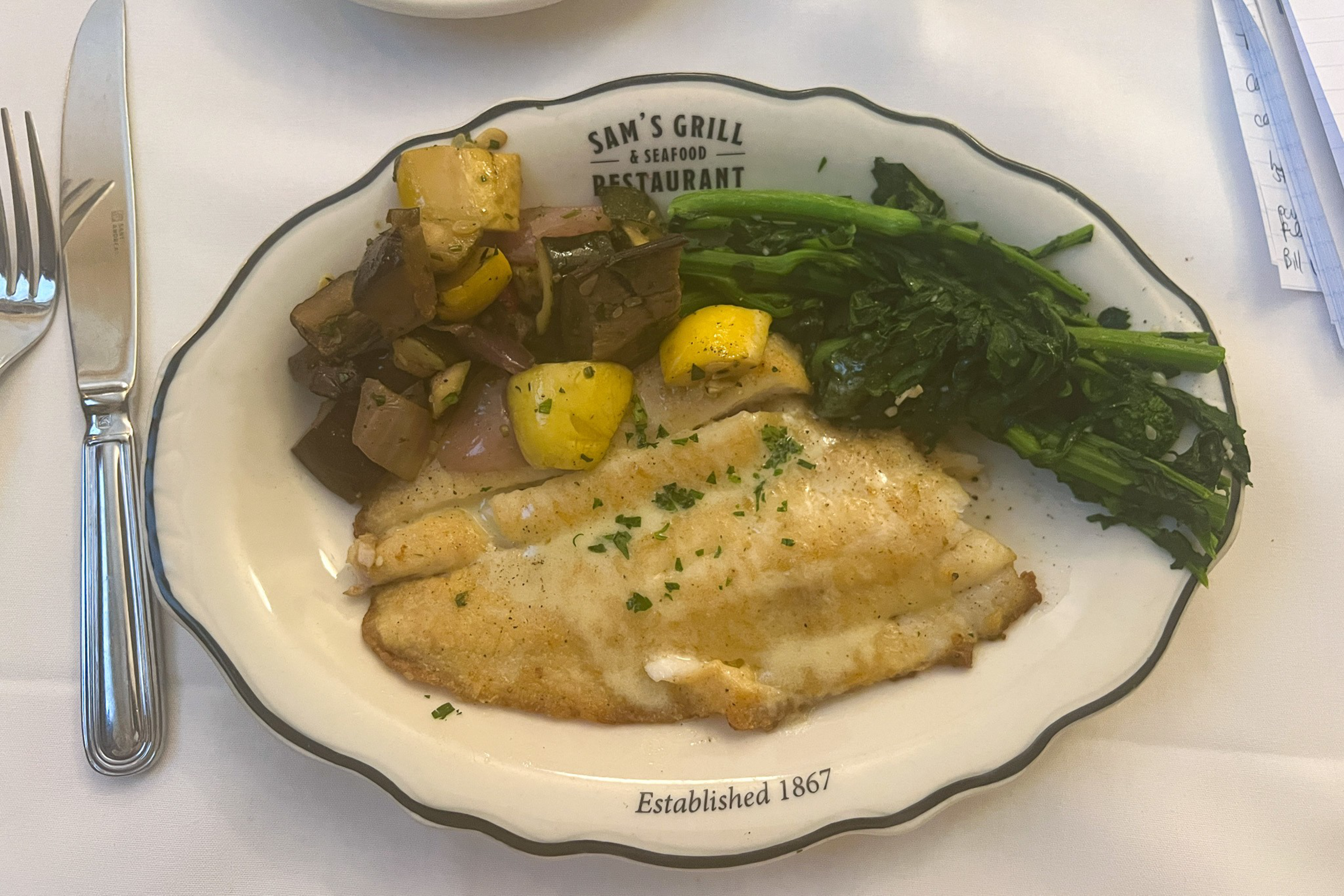 A plate of grilled white fish garnished with herbs, roasted vegetables including squash and eggplant, and a side of steamed greens.