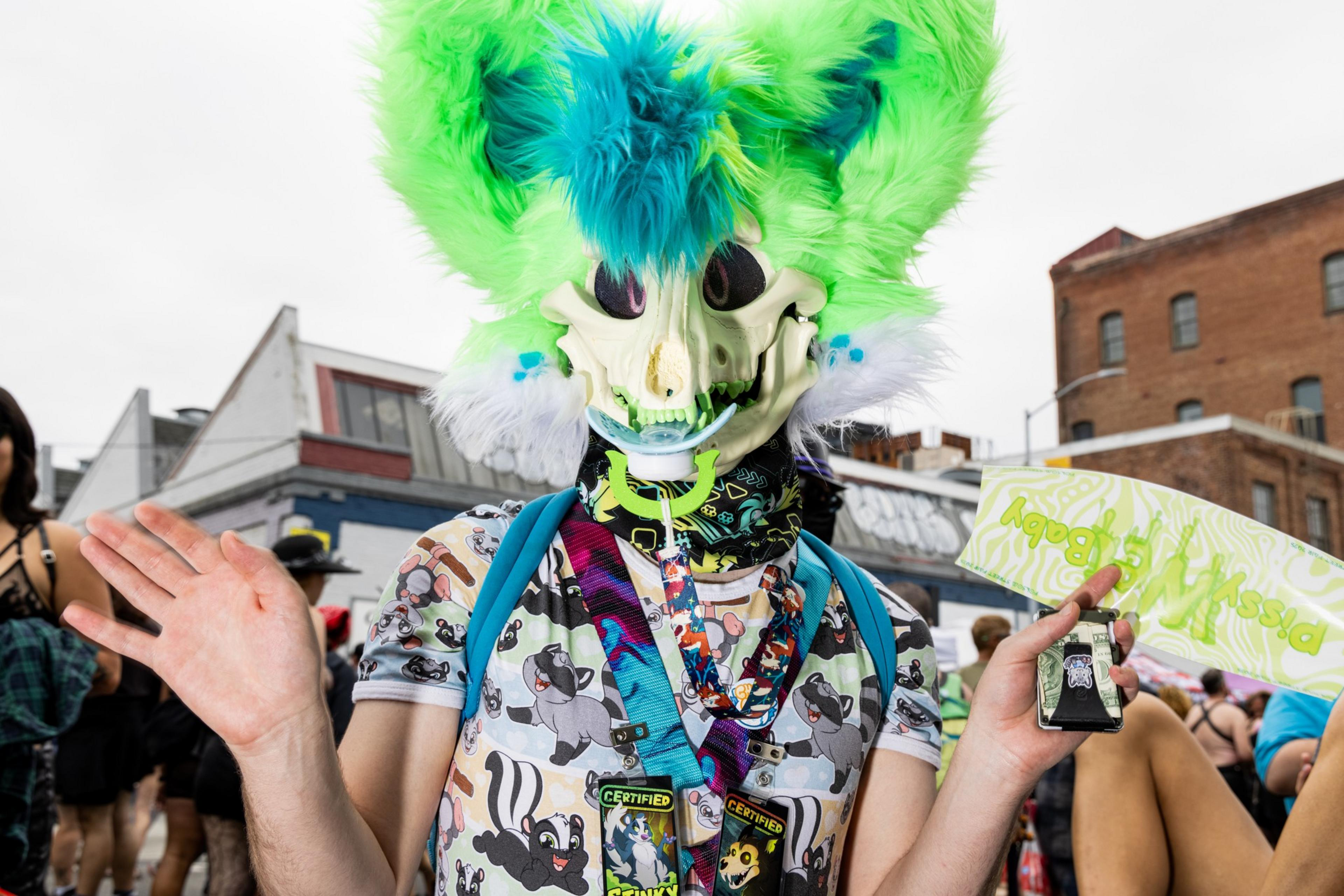 A person wearing a bright green and blue furry mask with a skull face, a dog-patterned shirt, and lanyards, waves their hands at an outdoor event.