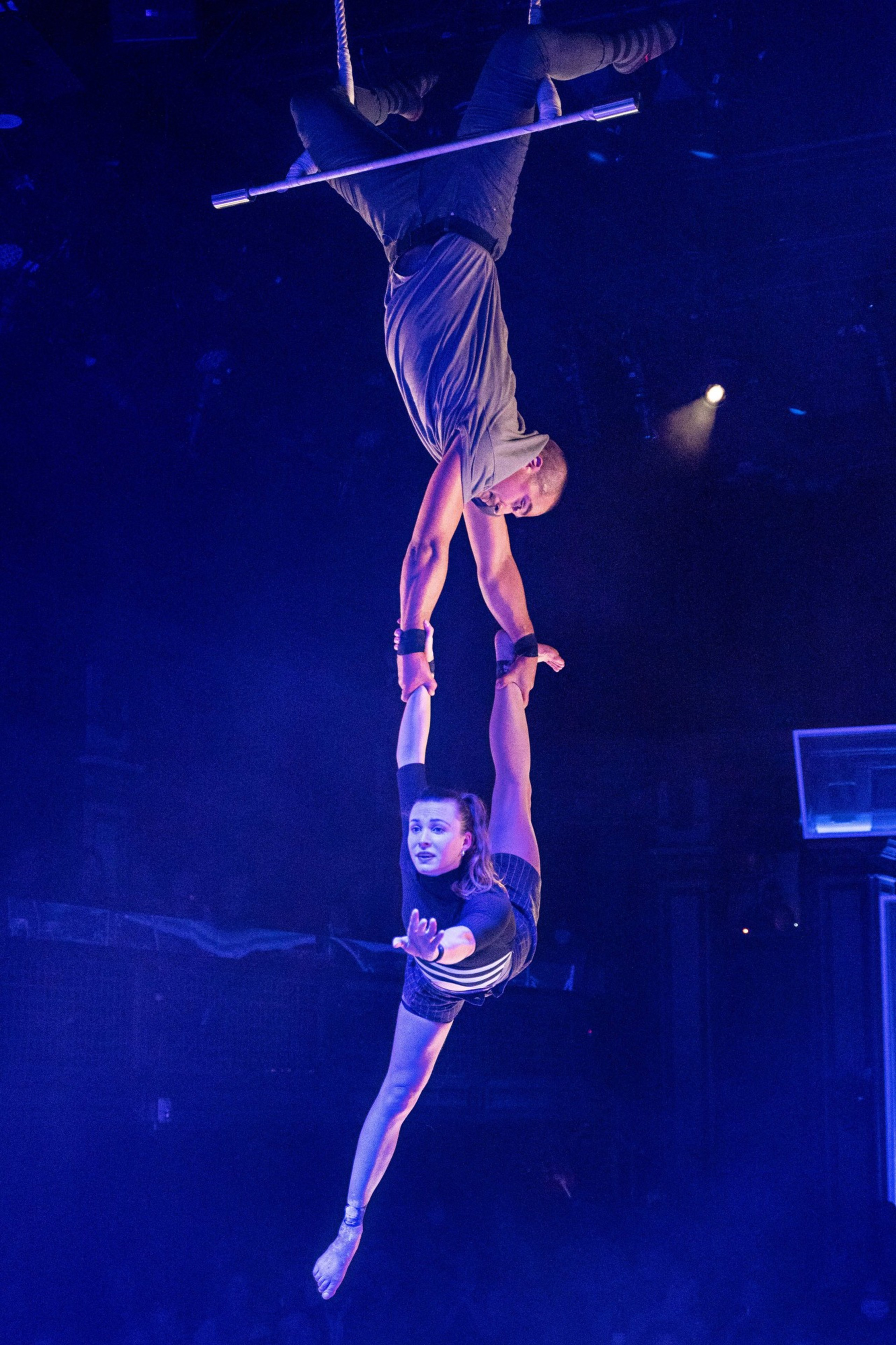 Two acrobats perform on a trapeze. One hangs upside down holding the other's hand, who is extended horizontally with one arm outstretched.