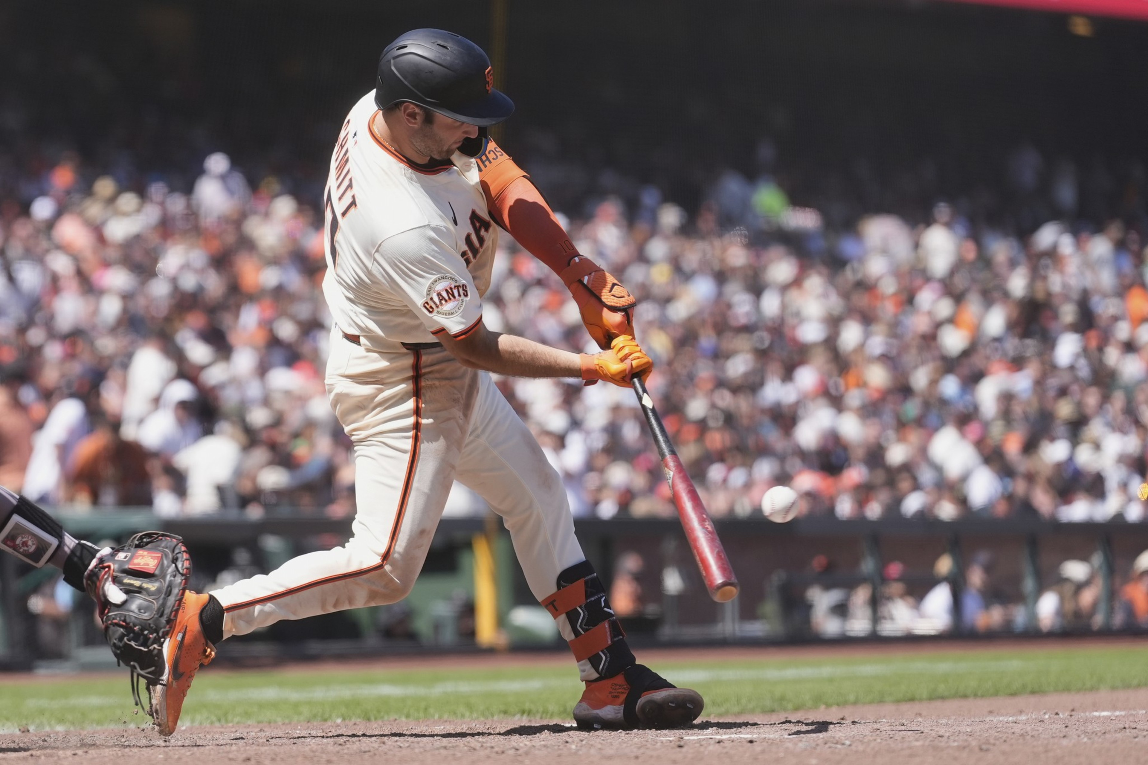 A baseball player in a white uniform is swinging a bat at a ball. The crowd in the stands is blurred in the background, and the player wears a helmet and orange gloves.