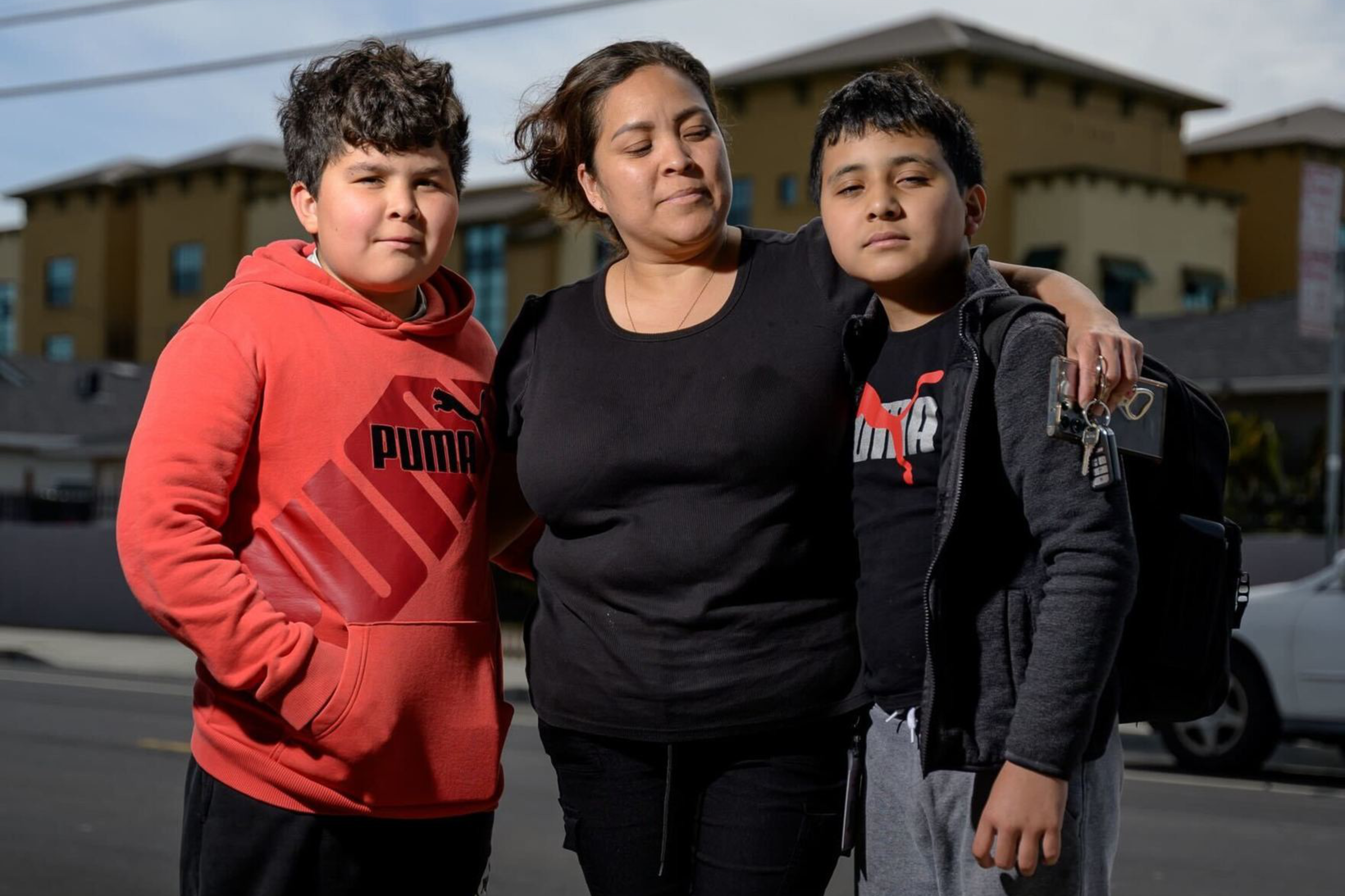 A woman stands on a sidewalk with her arms around two boys. One wears a red hoodie; the other has a backpack. They are in front of a building under a cloudy sky.