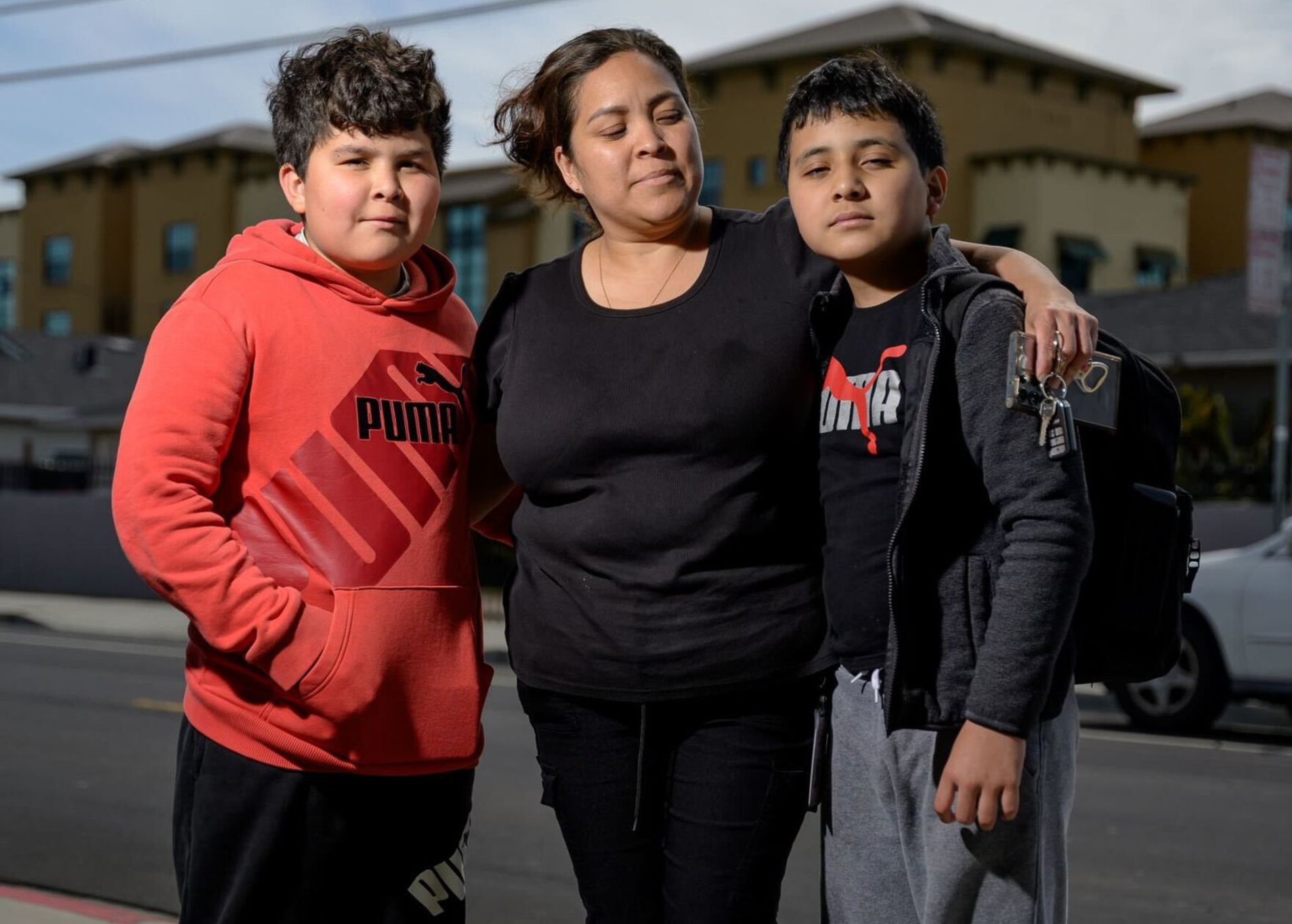 A woman stands on a sidewalk with her arms around two boys. One wears a red hoodie; the other has a backpack. They are in front of a building under a cloudy sky.