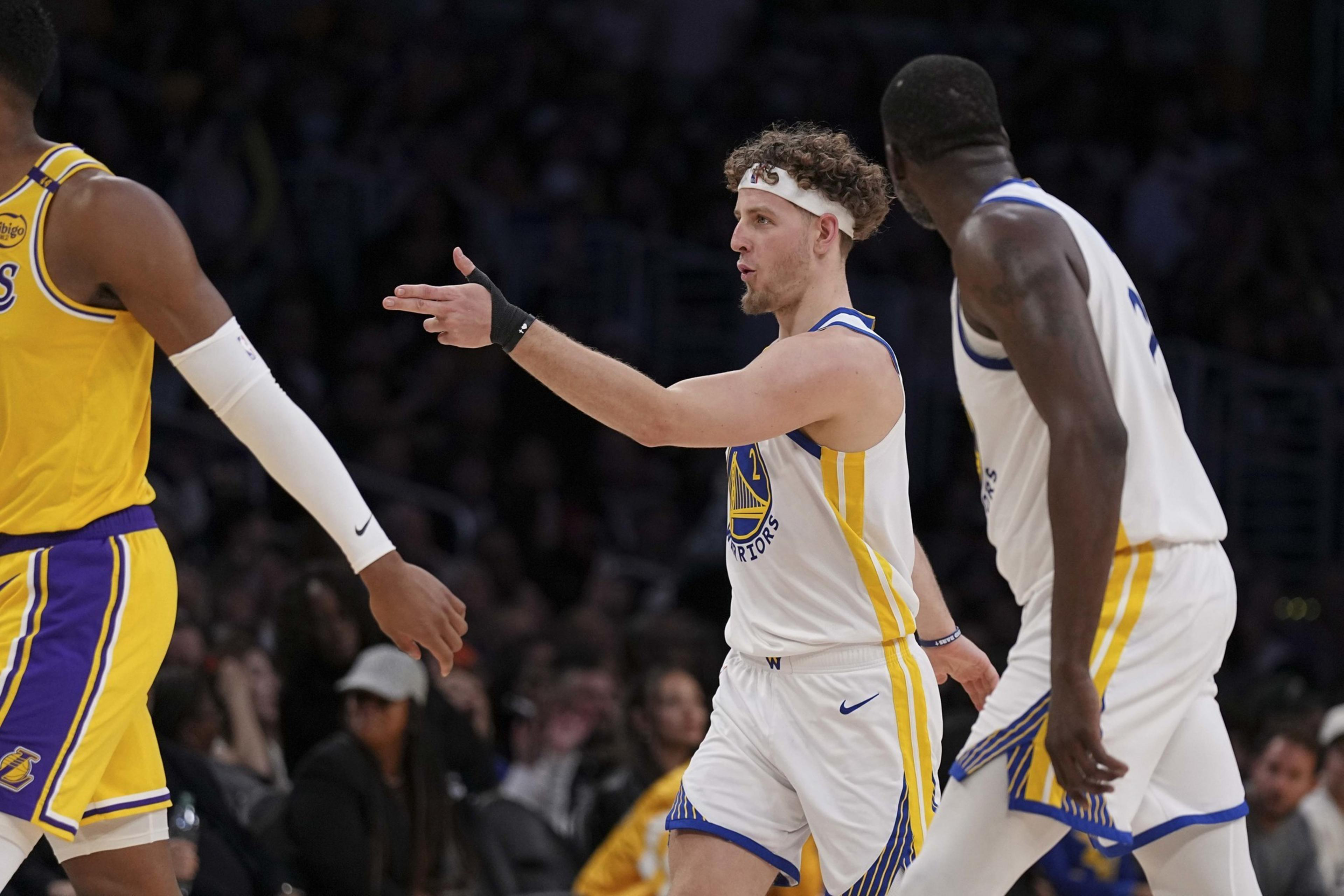 Two basketball players in white jerseys are interacting on the court, one gestures with his hand. A player in a yellow jersey walks by. The crowd is in the background.