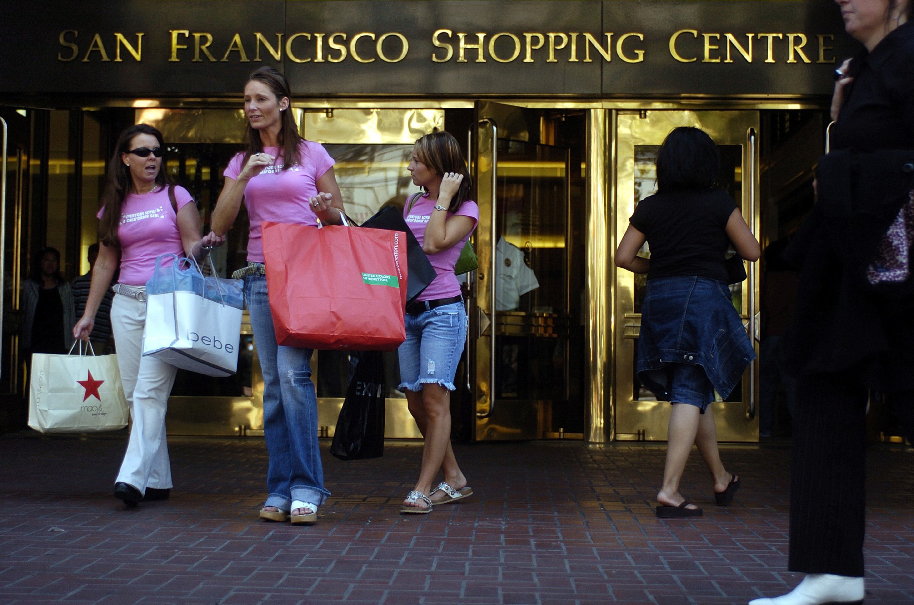 Three women in pink tops and jeans exit a shopping center with bags; one woman talks on a phone.