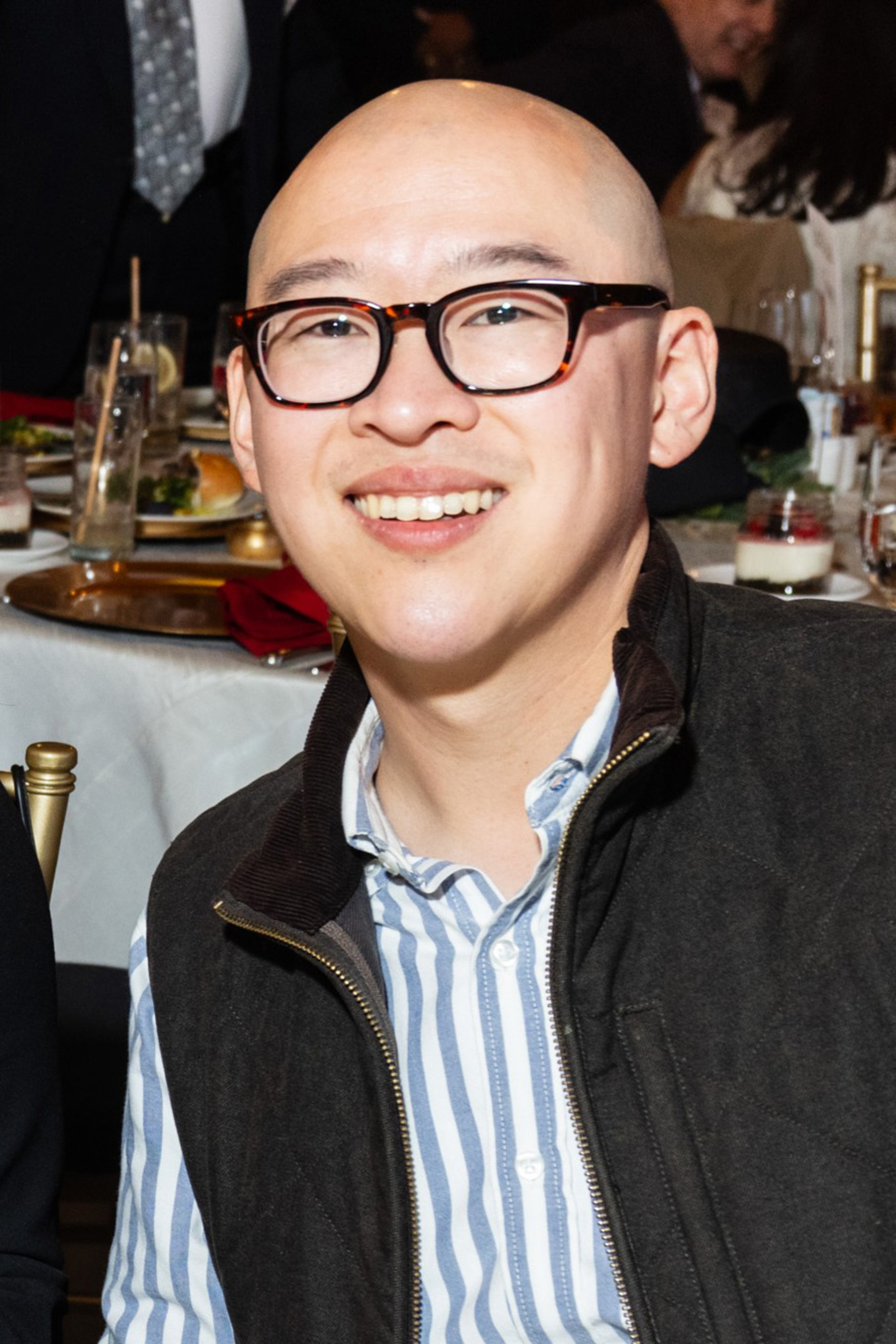 A smiling bald man with glasses wears a striped shirt and dark vest ...