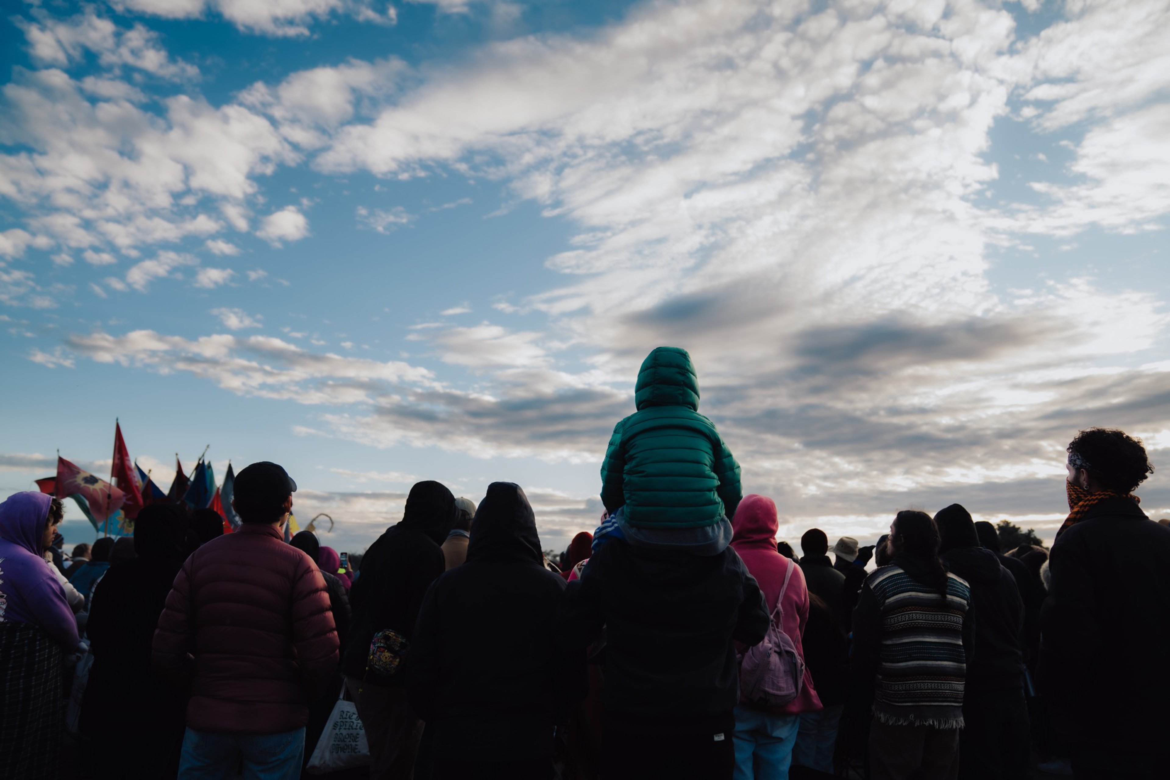 A crowd of people dressed in jackets and hoodies watch a cloudy sky, with a child in a turquoise jacket sitting on an adult’s shoulders.